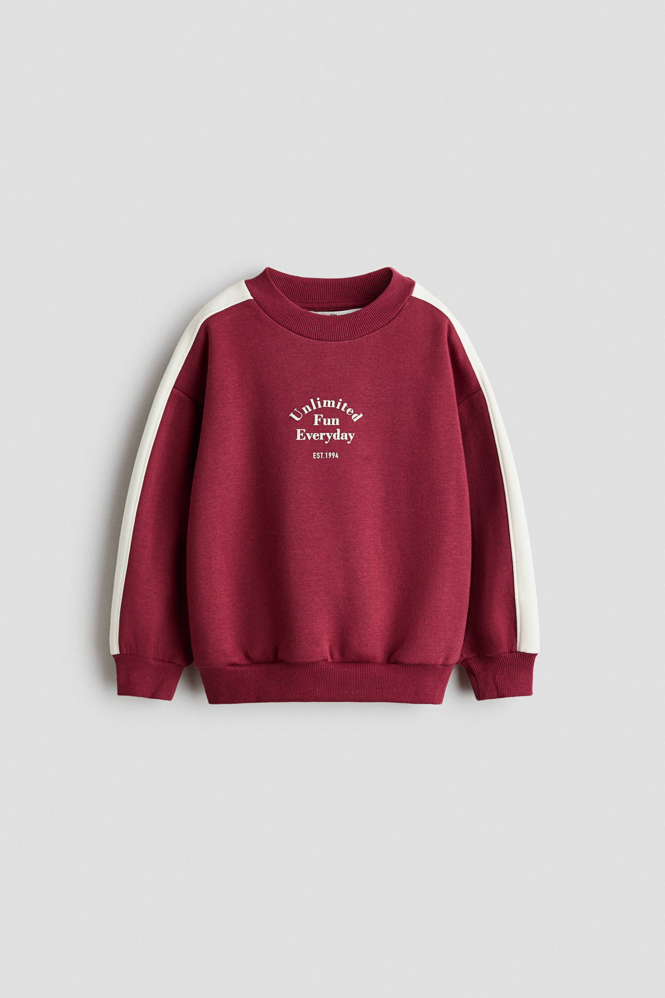 Xem ảnh lớn hơn: Một chiếc áo sweatshirt dáng rộng màu đỏ burgundy đang được chụp chính diện, nổi bật với cổ tròn bo rib, vai rơi và bo tay áo cùng gấu áo. Trên áo có chi tiết một dải sọc trắng tương phản chạy dọc mỗi tay áo và dòng chữ trắng ở giữa ngực ghi “Unlimited Fun Everyday EST.1994”.