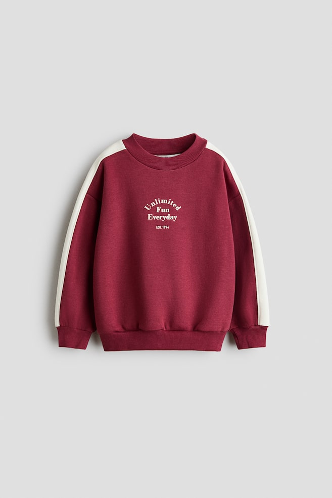 Una sudadera holgada color burdeos vista de frente, con cuello redondo rematado en canalÃ©, hombros caÃ­dos y puÃ±os y bajo de canalÃ©. La sudadera presenta una franja blanca en contraste que recorre cada manga y texto blanco en el centro del pecho que dice "Unlimited Fun Everyday EST.1994". - 1