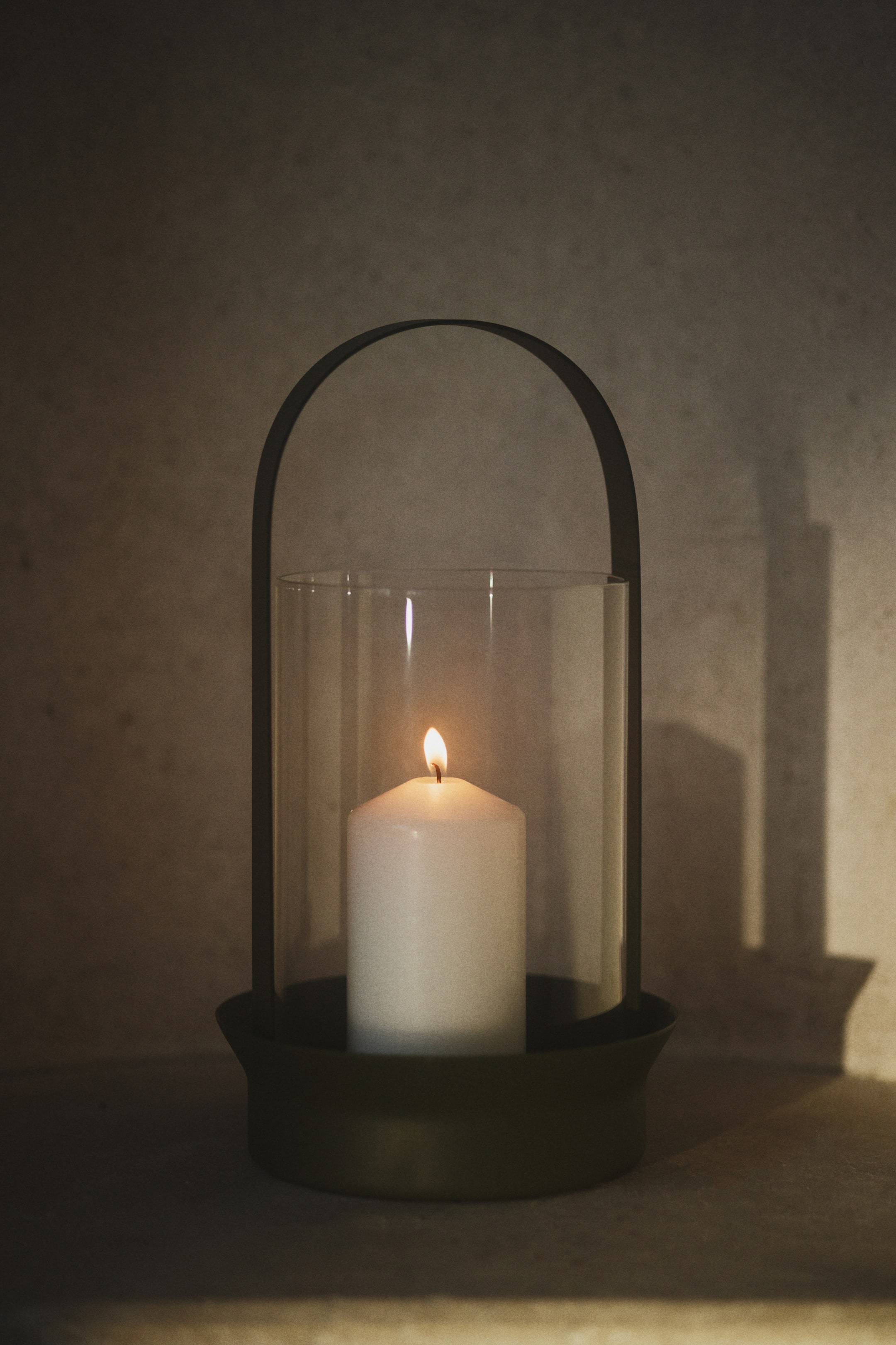 View larger image: A candle lantern featuring a dark olive green round base, a clear cylindrical glass wind guard, and a dark metal arched handle. A white pillar candle is burning brightly inside the glass.