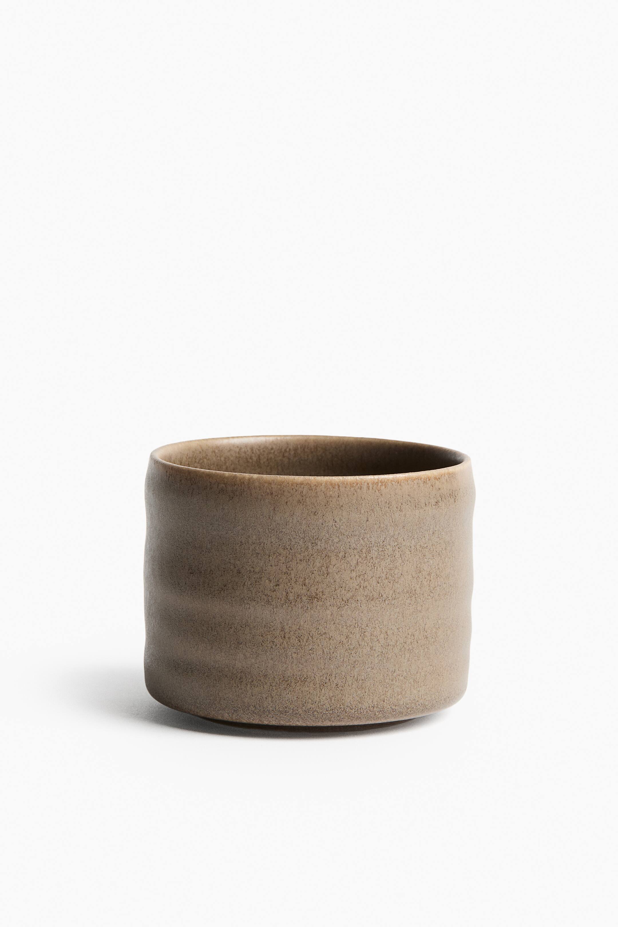 Grotere afbeelding bekijken: Een stenen mok staat recht van voren, met een zacht gegroefde vorm en subtiele horizontale ribbels rond het onderste deel. Het stuk heeft een matte, reactieve glazuurlaag in gevarieerde tinten beige en bruin, met een gespikkelde, textuurrijke uitstraling.