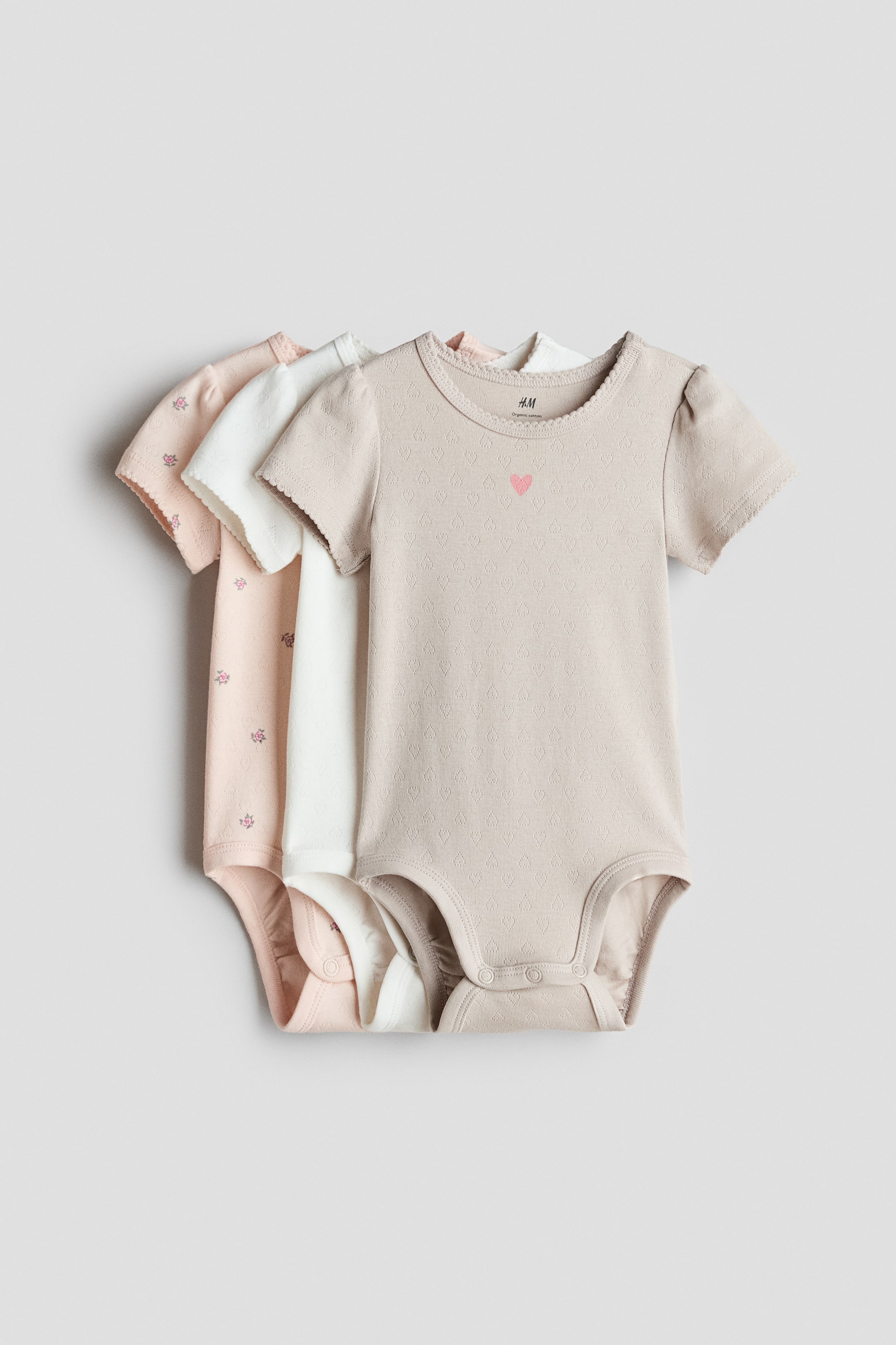 View larger image: Three short-sleeved bodysuits are displayed from the front, layered together. The front beige cotton jersey bodysuit features a subtle heart pattern, a small pink embroidered heart, a round neckline with picot trim, a press-stud on one shoulder, short sleeves with picot trim, press-studs at the bottom, and gathered leg seams. A white bodysuit and a light pink floral-patterned bodysuit are visible behind it.