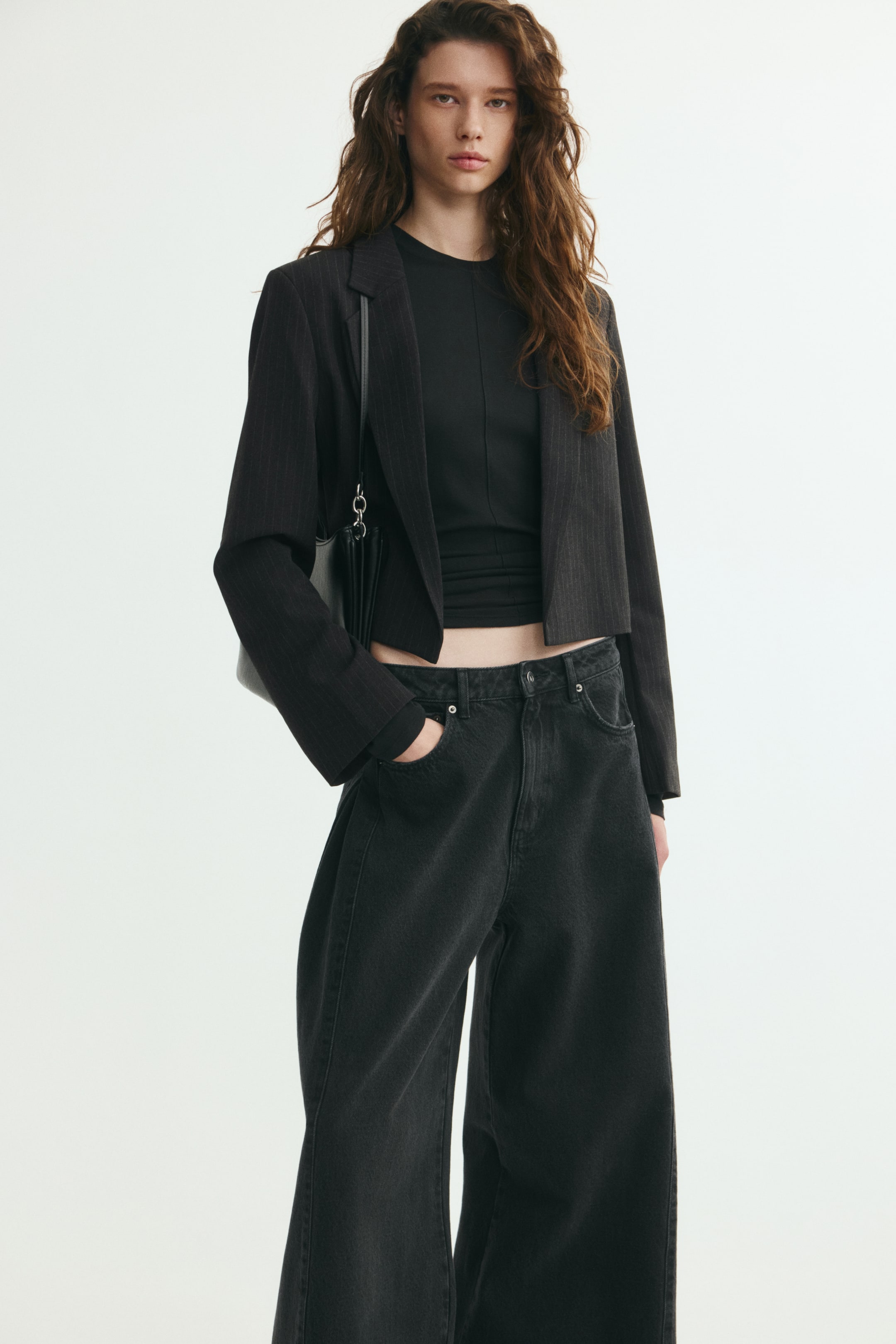 Vis større bilde: En cropped blazer i mørk grå teksturert twill bæres av modellen over en svart topp og stylet med mørke jeans med vide ben. Den åpen-front blazerjakken har strukturerte skulderputer og jakkeslag med notch, med en svart skulderveske båret under armen.