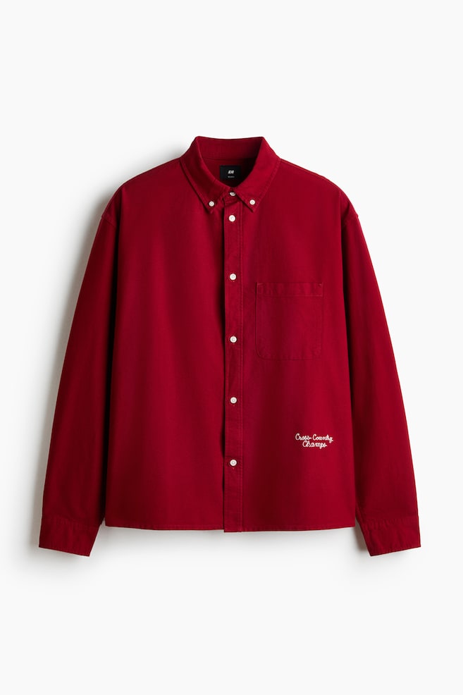 Une chemise rouge à manches longues est présentée de face, avec un col boutonné, une patte de boutonnage complète avec des boutons blancs, et une poche plaquée sur la poitrine gauche. Un texte brodé en blanc, "Cross-Country Champs", est visible sur le bas avant droit. - 1