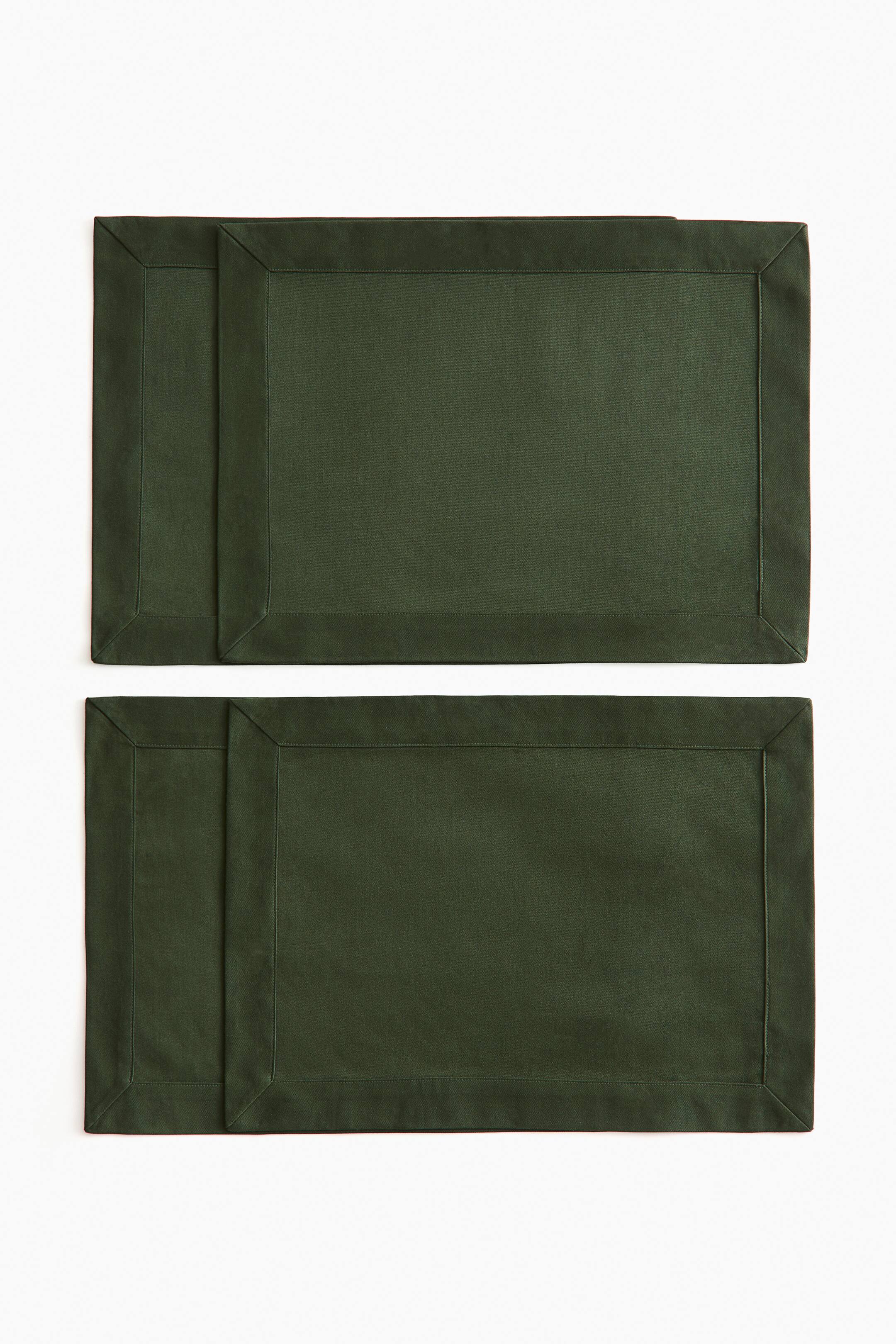 View larger image: Dark green cotton placemats are laid flat in two slightly overlapping pairs, showcasing their rectangular design from a front overhead angle. Each placemat features a wide stitched border with folded hems and mitred corners, highlighting a clean and structured finish.