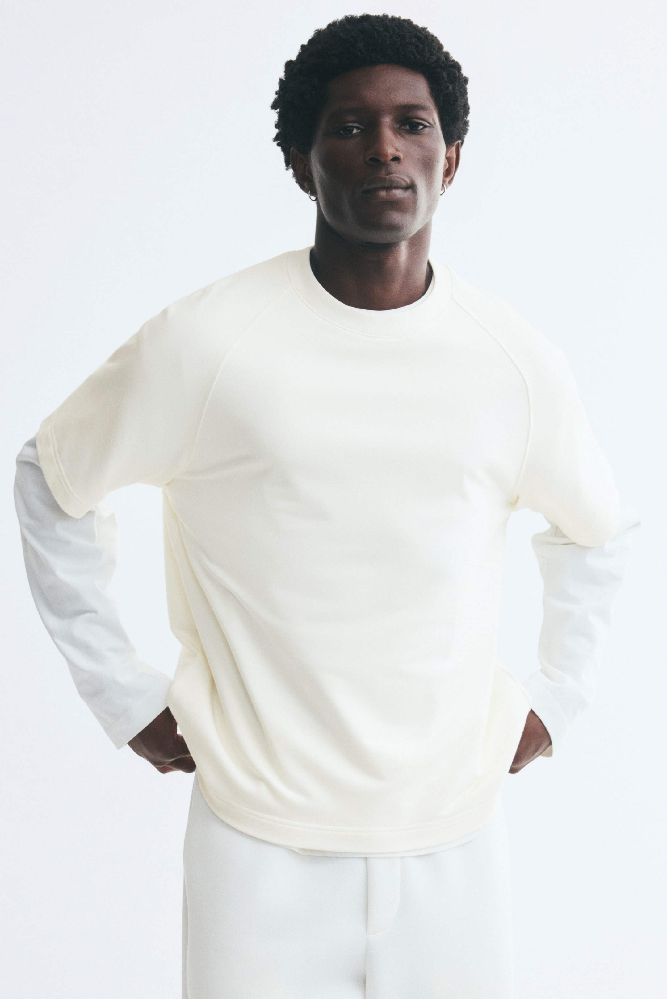 Voir une image plus grande: Un t-shirt en jersey épais de couleur crème, avec col rond côtelé et manches raglan courtes, est porté par le mannequin par-dessus une chemise blanche à manches longues. Le t-shirt à coupe décontractée présente un ourlet droit, tandis que le mannequin repose ses mains près de ses hanches.