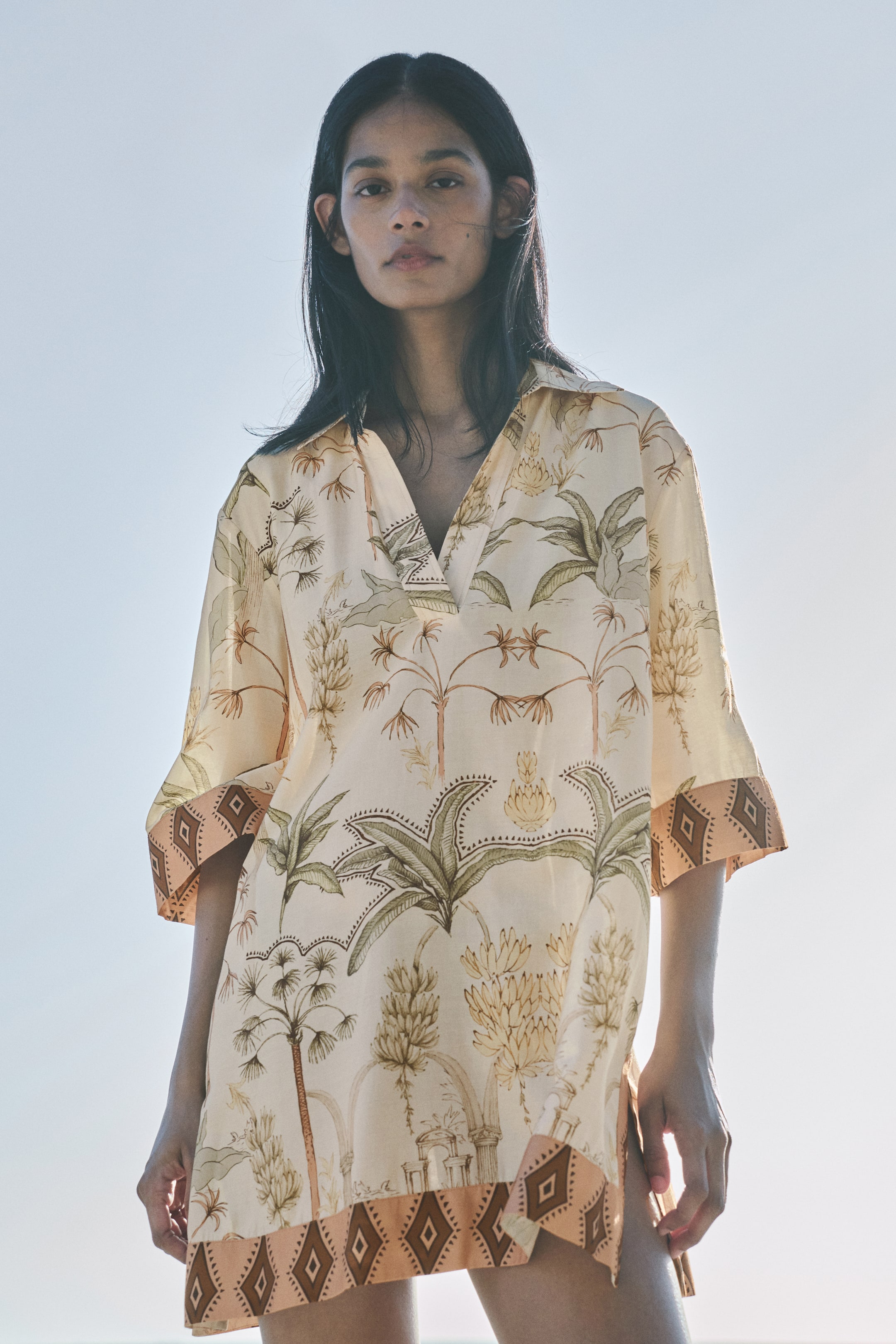 View larger image: A cream tunic-style top featuring a botanical print of palm trees and banana plants, with a v-neck and a spread collar. The wide, three-quarter length sleeves and straight hem showcase a peach and dark brown patterned band with diamond shapes.