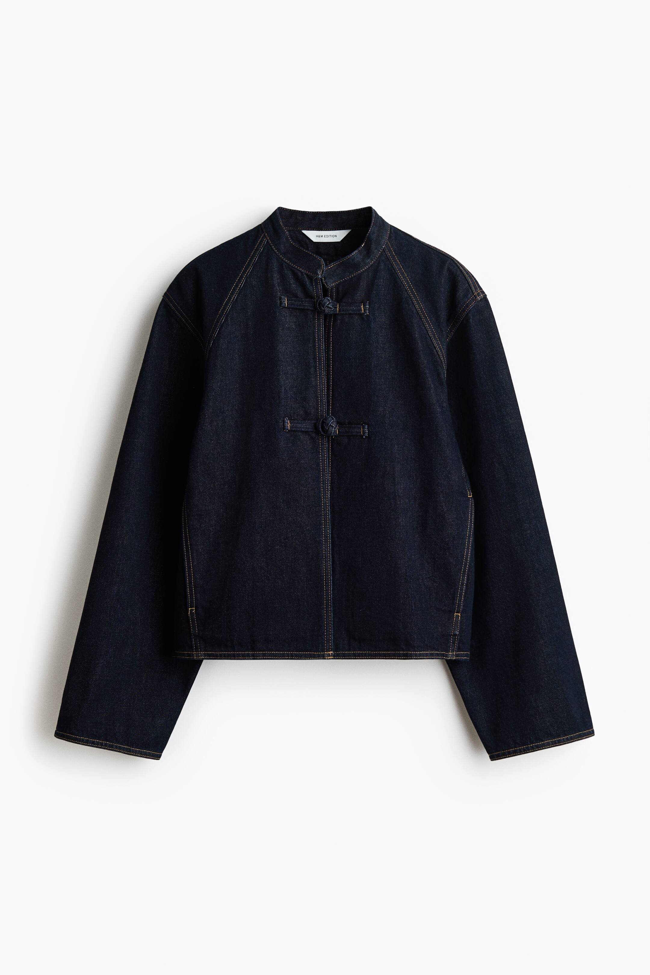 View larger image: The front view showcases a dark indigo denim jacket. It features a stand-up collar with three frog closures along the front placket, and the raglan sleeves and seams are detailed with contrast light brown stitching.