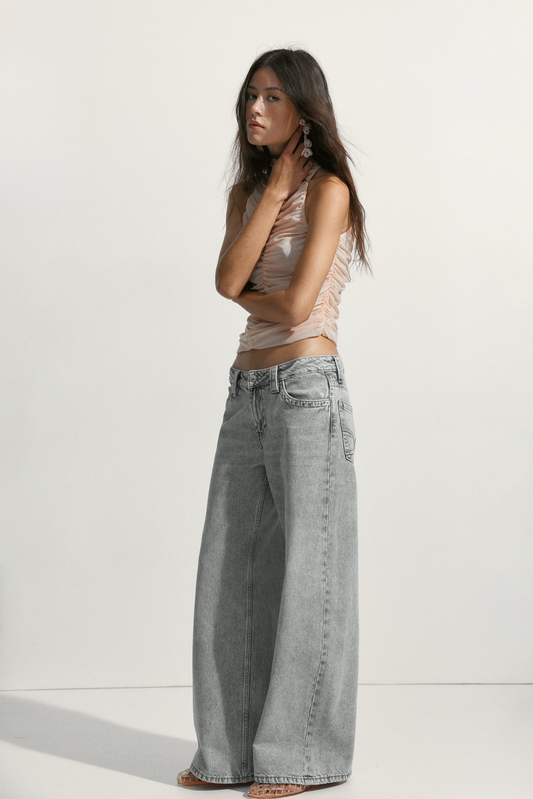 הצגת תמונה גדולה יותר: חולצת קרופ ללא שרוולים בצבע ורוד בהיר עם כיווצים, מדגישה בד מרקם קליל ושקוף במעט וגזרה צמודה. היא משתלבת עם ג'ינס אפור בהיר בגזרה גבוהה ורחבה, המציג סילואט רפוי באורך מלא עם שטיפה דהויה.