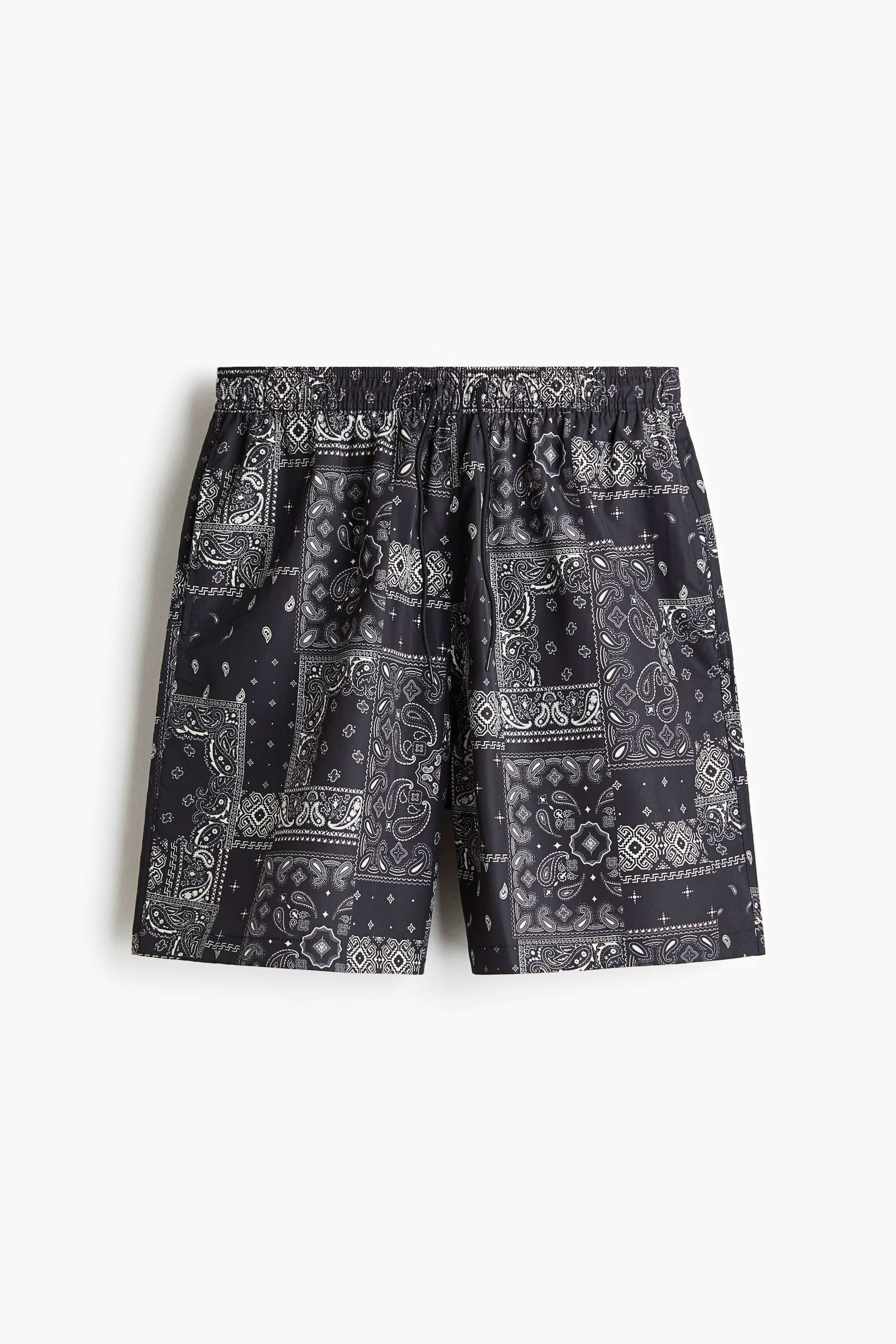Agrandir l'image: Les shorts arborent un motif patchwork bandana noir et blanc. Ils sont conçus avec une taille élastiquée et un cordon de serrage visible, offrant une coupe décontractée mi-cuisse.