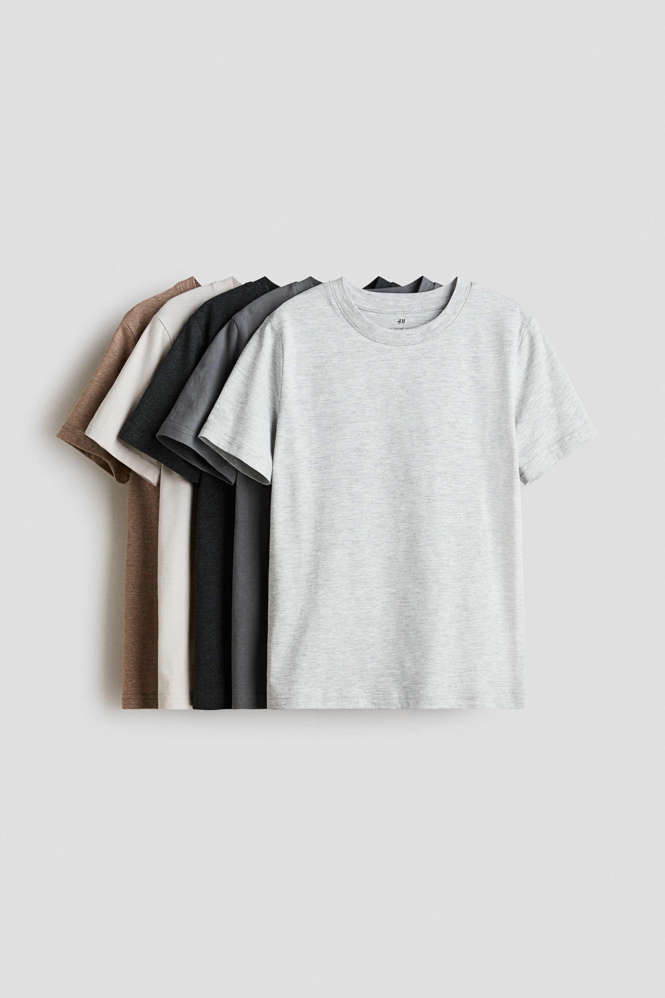 Ampliar la imagen: Se presenta una camiseta de jersey de algodón suave en color gris claro, con cuello redondo y dobladillo recto. Detrás de ella, se disponen en forma de abanico cuatro camisetas adicionales en colores gris oscuro, negro, beige y marrón.