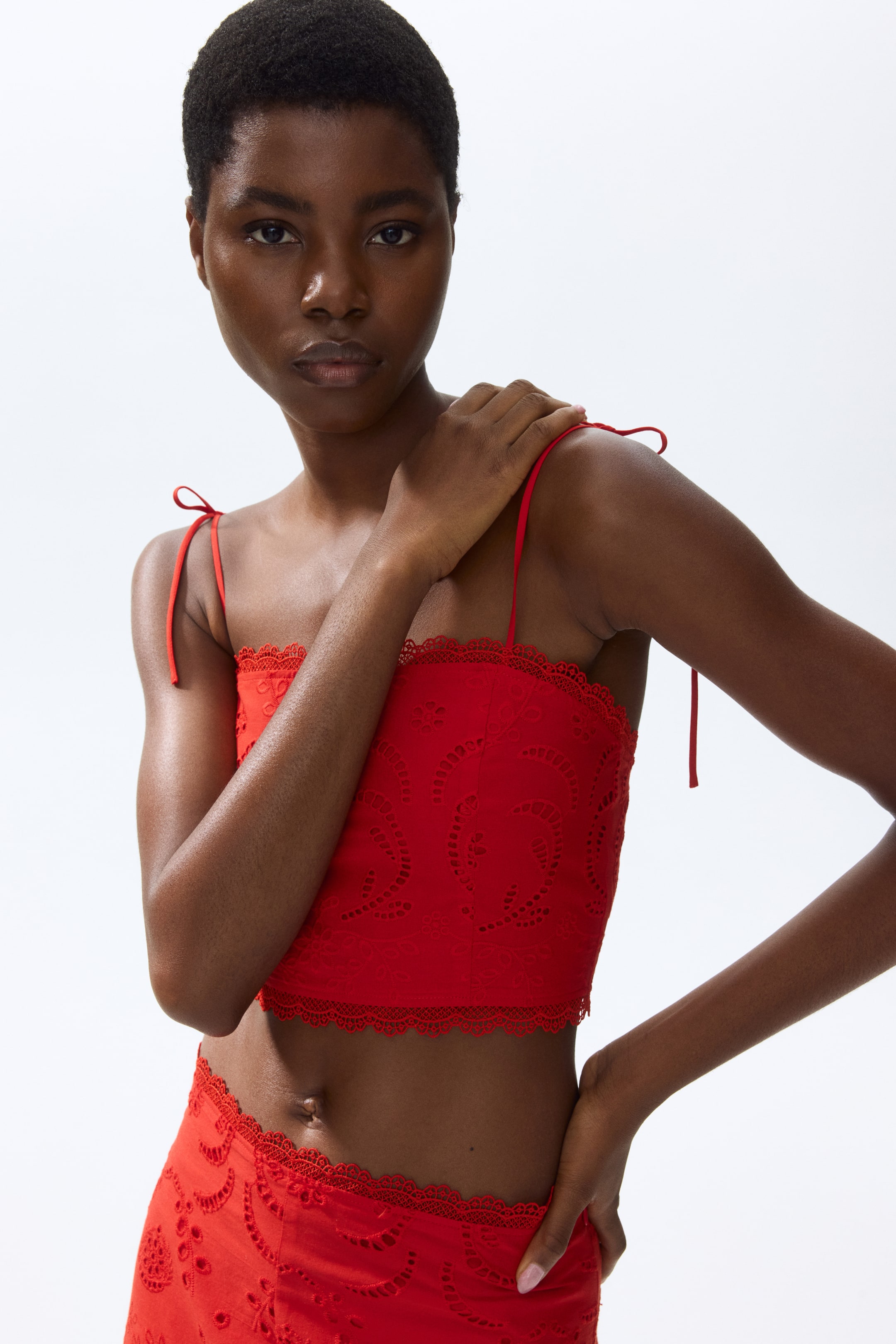 View larger image: A red eyelet embroidery cropped top and matching bottom featuring circular and floral patterns. The top is designed with thin, tie-up spaghetti straps, one tied into a bow on the left shoulder, and both pieces showcase scalloped lace trim along their edges.