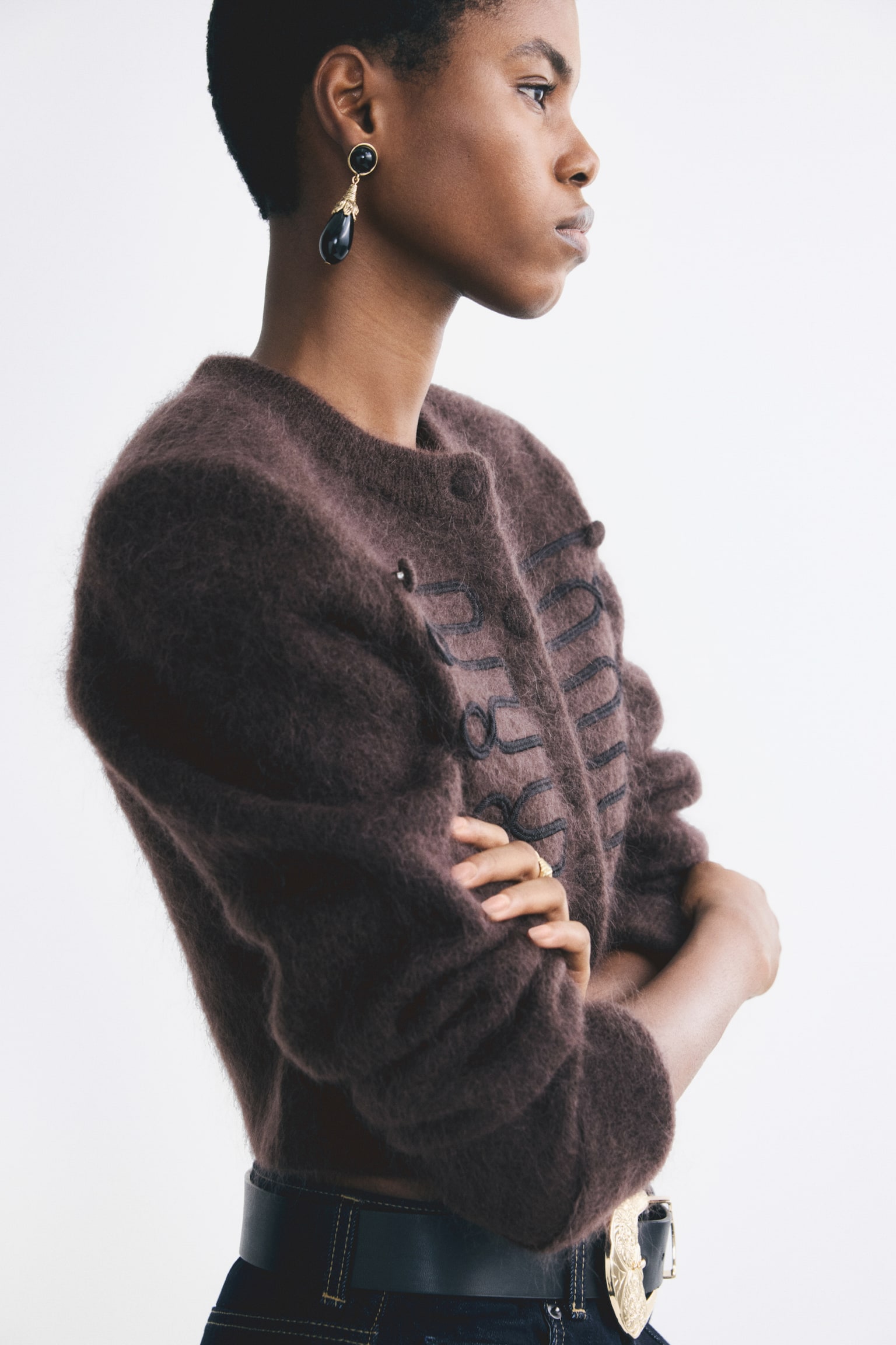 A brown mohair blend shoulder pad cardigan is being worn by the model, who is standing in profile with arms crossed. The cardigan features a fuzzy, textured knit, structured shoulders, and black embroidered detailing along the front. - 1