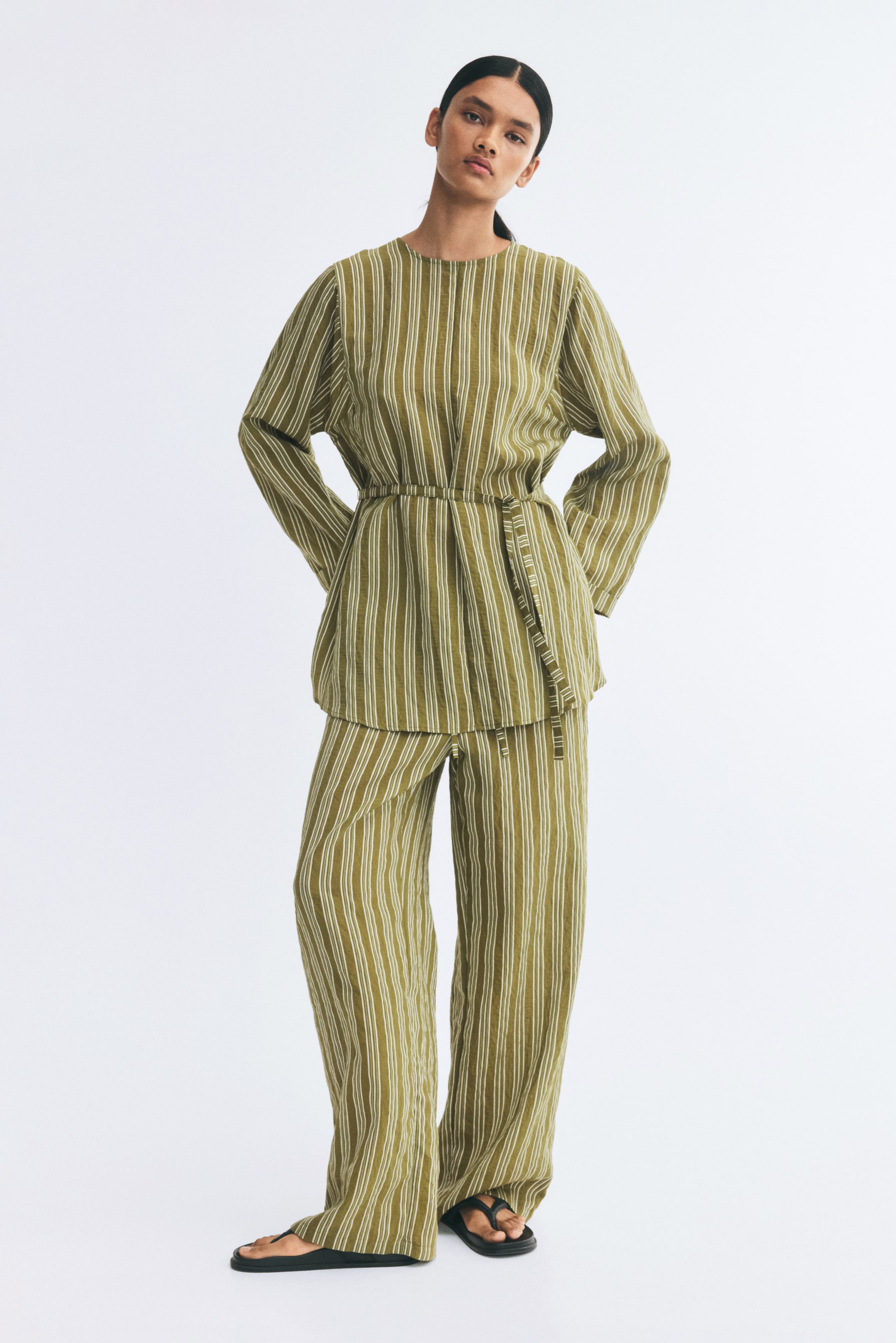 View larger image: Loose-fit, straight-leg pants featuring a vertical olive green and white striped pattern are worn by the model. The pants are styled with a matching long-sleeved striped tunic, belted at the waist, and black sandals.