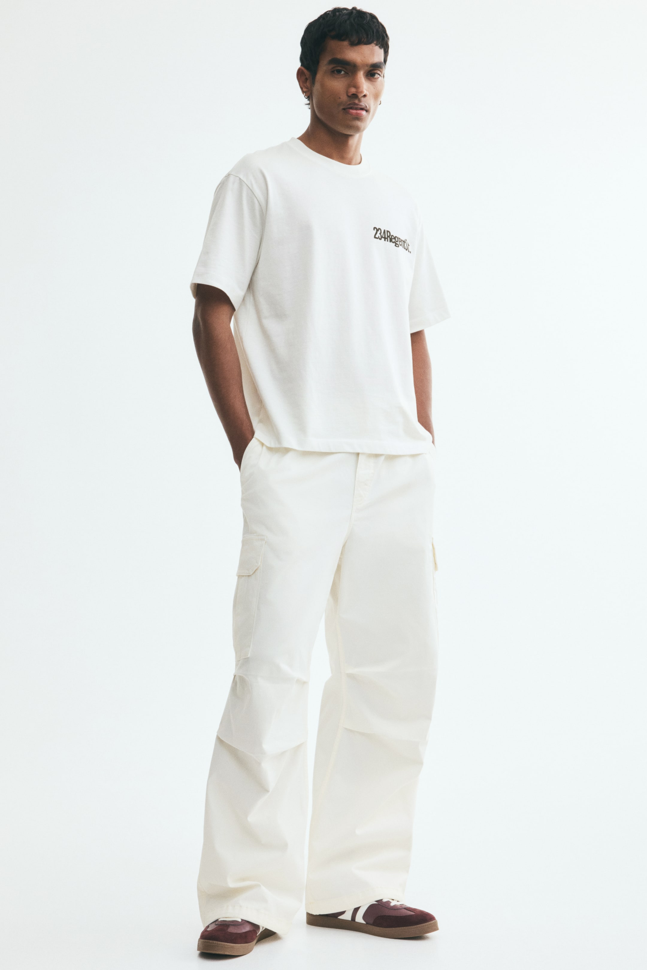 View larger image: The model is wearing loose-fit cargo trousers featuring a covered elastic waistband and a drawstring at the waist, diagonal side pockets, patch leg pockets with flaps, pleats at the knees, and drawstrings at the hems. The model is placing their hands into the diagonal side pockets.