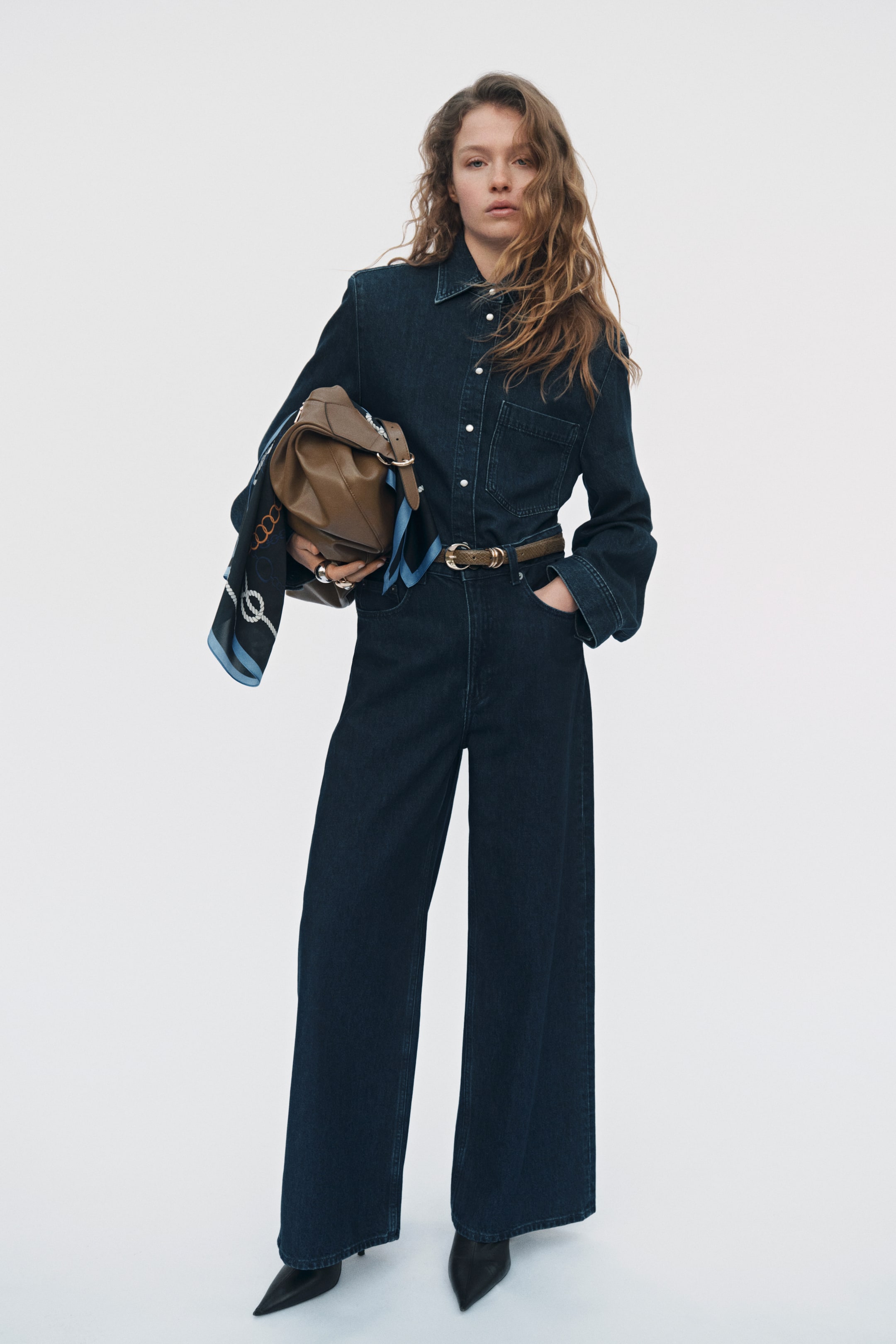 Voir une image plus grande: Une combinaison en denim délavé foncé à l'avant présente un haut boutonné à col avec des manches longues, de larges poignets et une poche poitrine. La combinaison continue avec un pantalon à jambes larges, et une fine ceinture à motifs ceinture la taille.