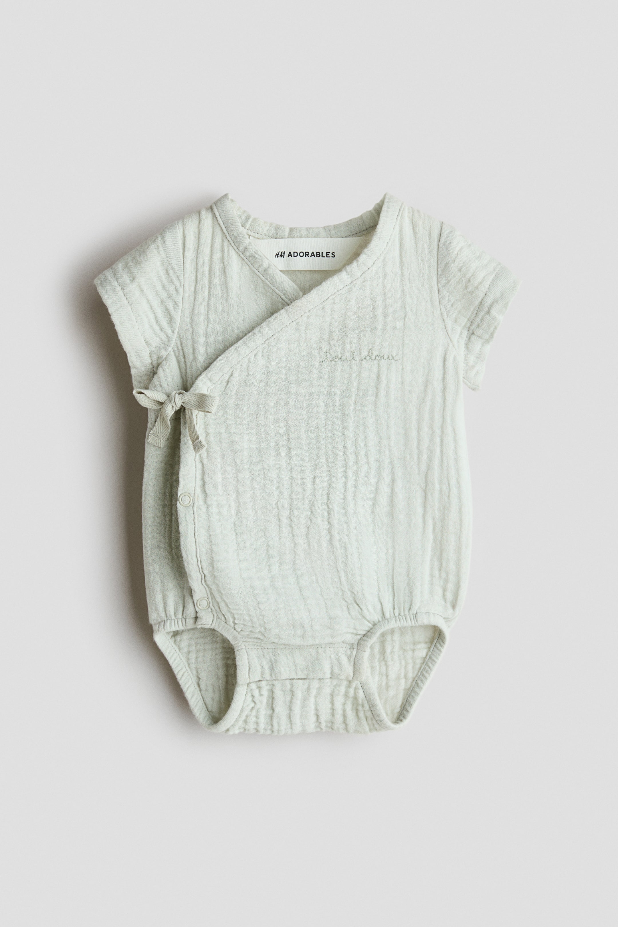 Visa större bild: En ljusgrön babyromper med en krinklad struktur och en kimono-stil omlottfront, som fästs med ett band på vänster sida och med tryckknappar längs kroppen. Den är designad med korta ärmar, elastiska benslut och "tout doux" broderat på höger bröst.