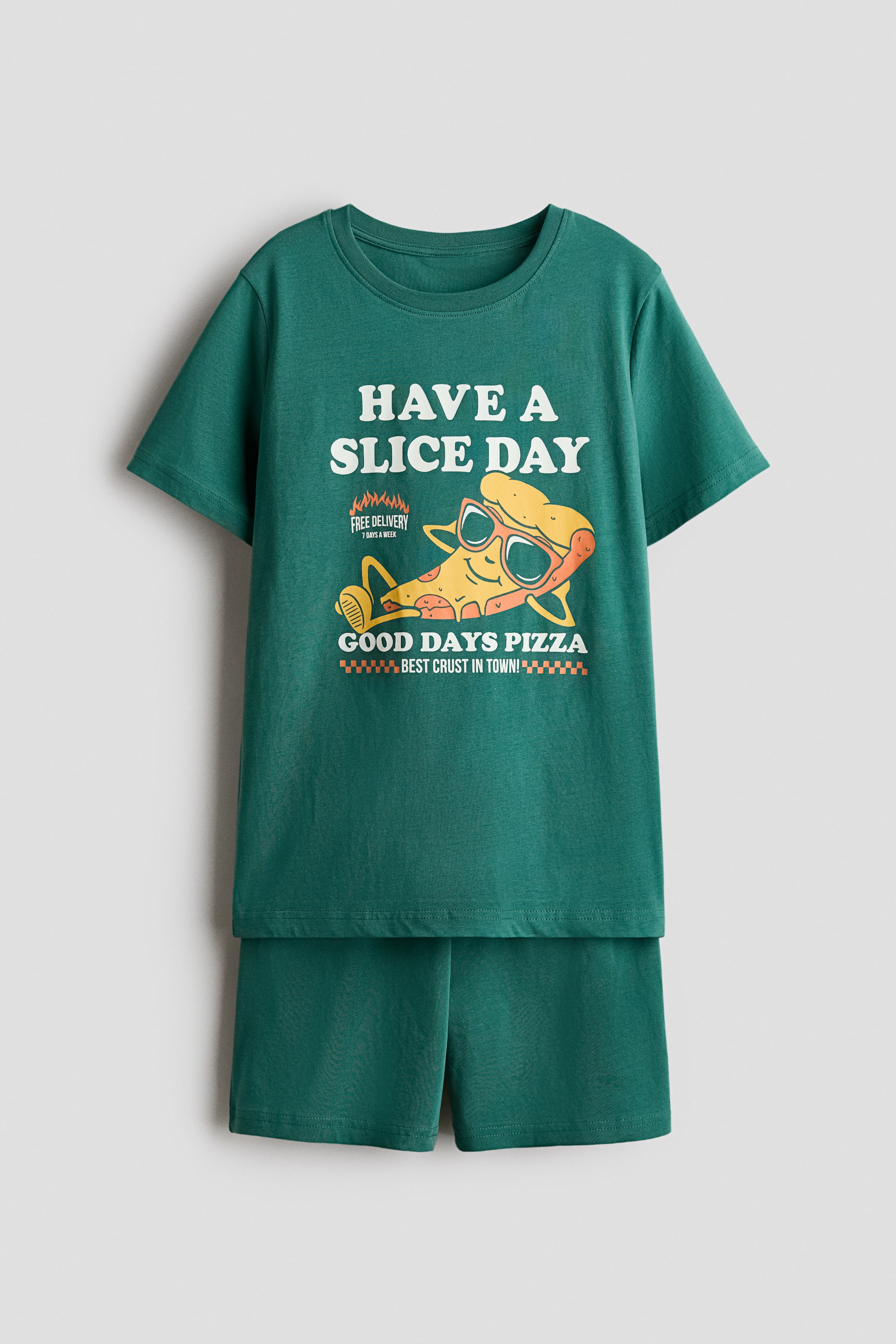 Grotere afbeelding bekijken: Pyjama met een groene T-shirt met korte mouwen, voorzien van ribbels rondom de halslijn en een "HAVE A SLICE DAY" pizza-graphic aan de voorkant, en bijpassende groene shorts met een elastische tailleband, worden van voren gepresenteerd.