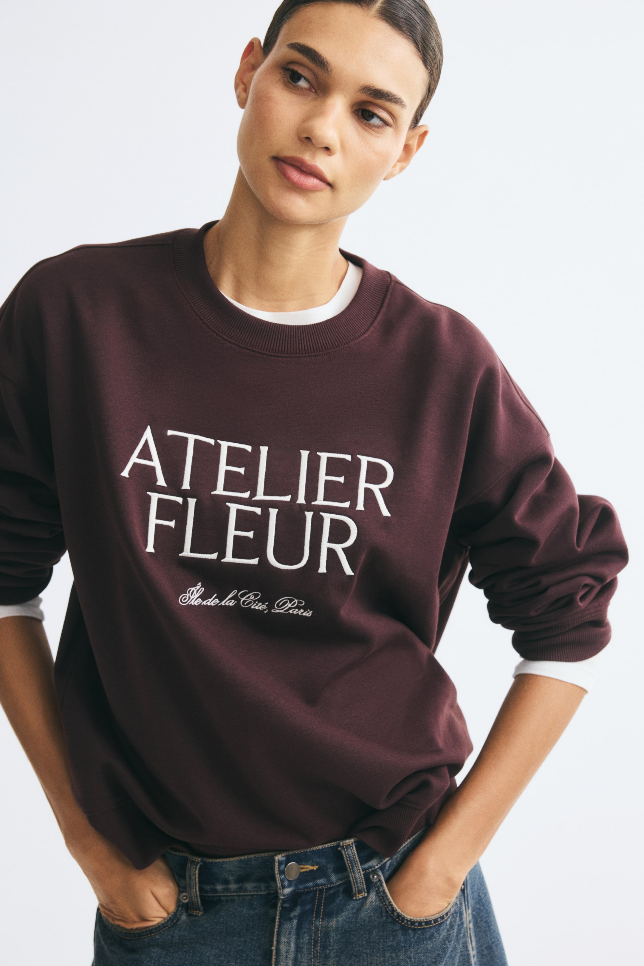 View larger image: A dark burgundy oversized sweatshirt featuring white text reading "ATELIER FLEUR" on the front is worn by the model. The sweatshirt features a ribbed crew neckline and dropped shoulders, and is layered over a white t-shirt and styled with blue jeans.
