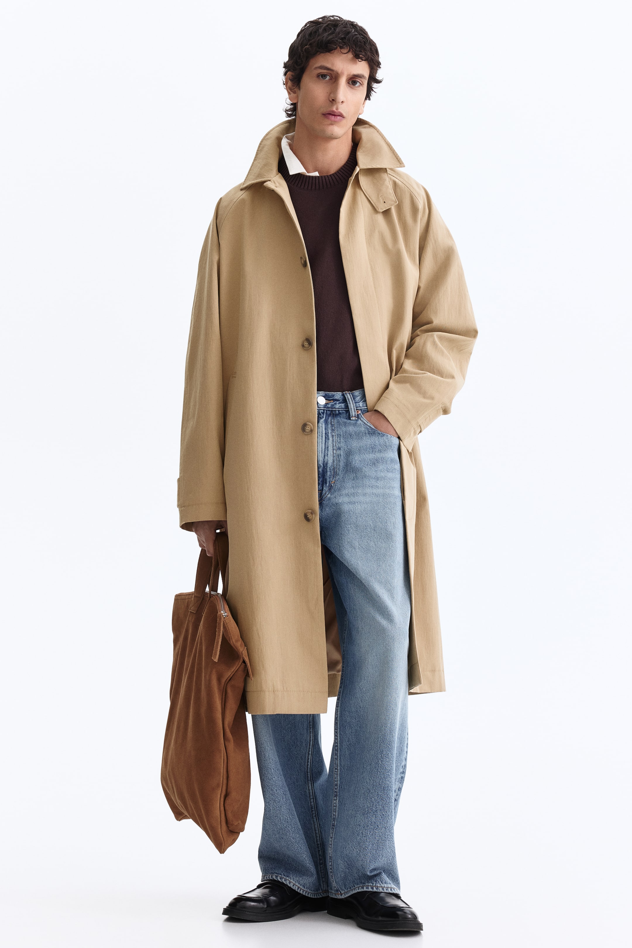 View larger image: The front-facing beige trench coat features a spread collar, raglan sleeves and a single-breasted button-front closure with three visible buttons, falling below the knee. It is worn open, revealing a brown crew-neck sweater layered over a white collared shirt.