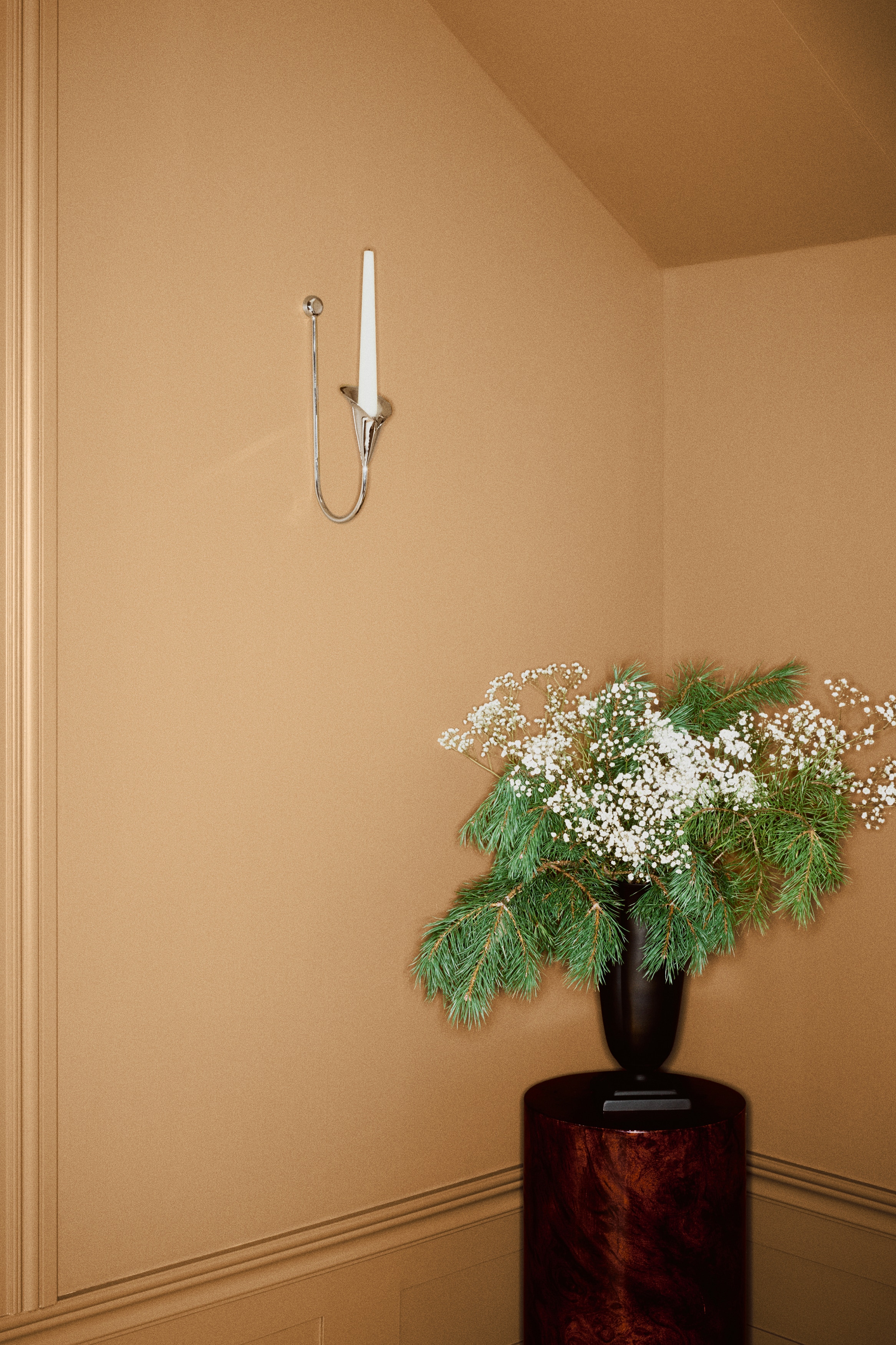 Een wandkandelaar van metaal in zilverkleur wordt frontaal getoond, met een lange, verticale stang met aan de bovenkant een bolvormige knop. De stang buigt aan de onderkant omhoog en eindigt in een hoogglanzende kaarsenhouder in de vorm van een calla lelie, met een reflecterende afwerking.
