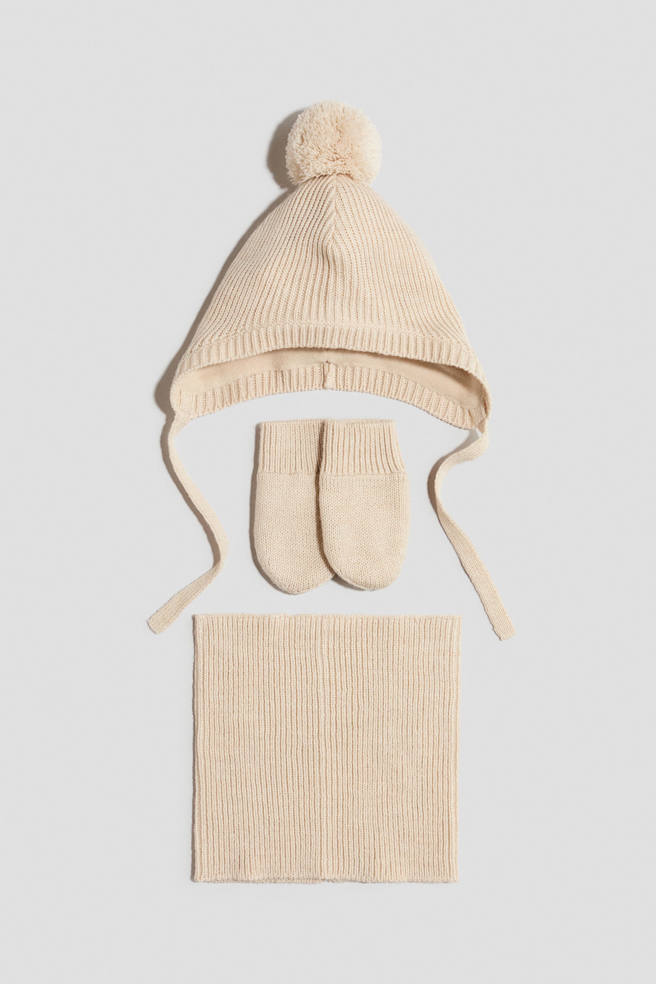 View larger image: A cream knitted winter set features a bonnet-style hat with a pom-pom, ear flaps, and ties, along with a pair of mittens and a rectangular scarf. All pieces display a vertical ribbed knit pattern, with the hat showing a soft lining at the brim and the mittens having ribbed cuffs.