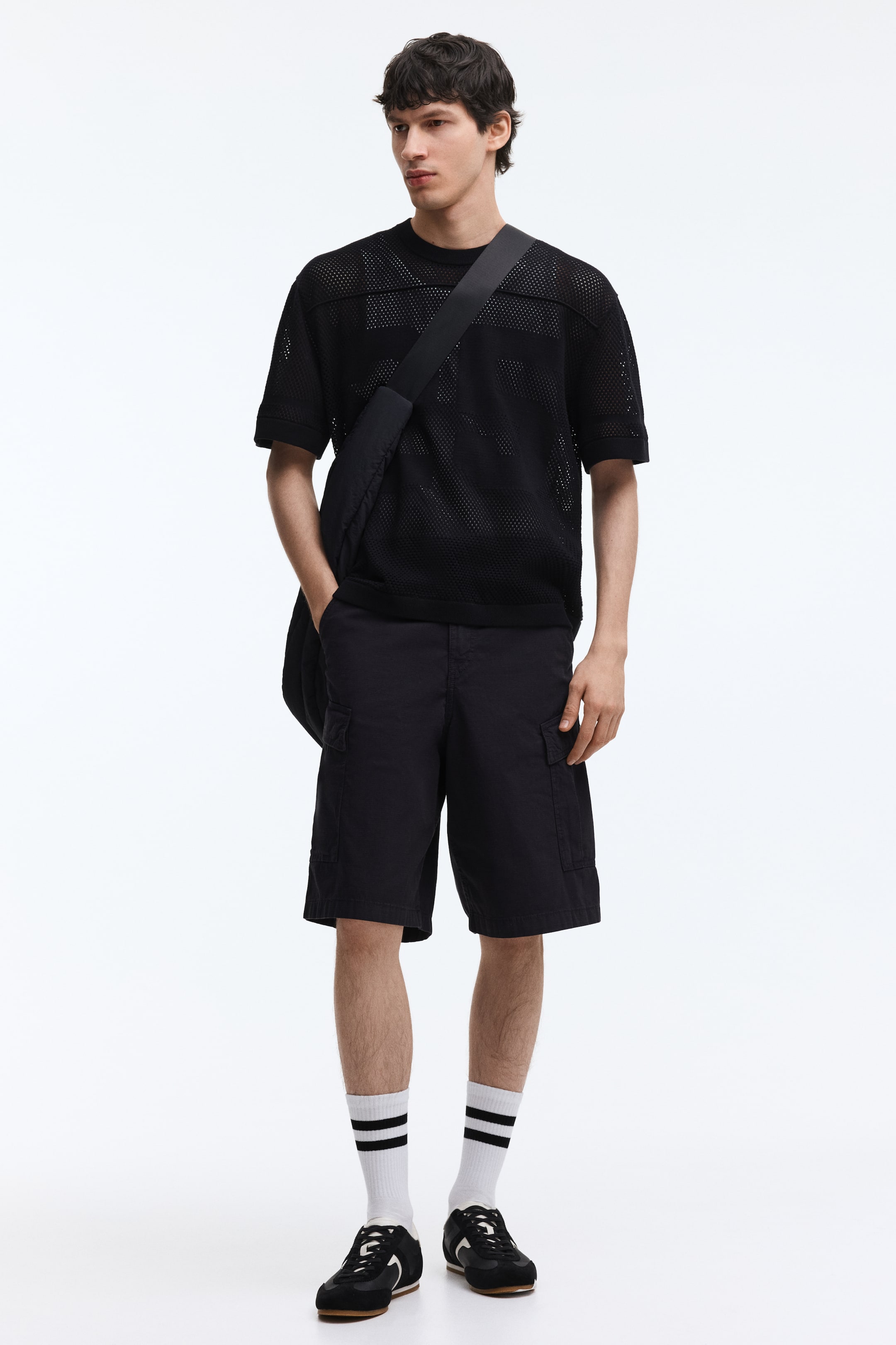 View larger image: A black mesh short-sleeved t-shirt with solid horizontal panels across the chest, ribbed cuffs, and a ribbed hem is paired with black cargo shorts featuring side flap pockets. A black cross-body bag with a wide strap is draped diagonally across the t-shirt.