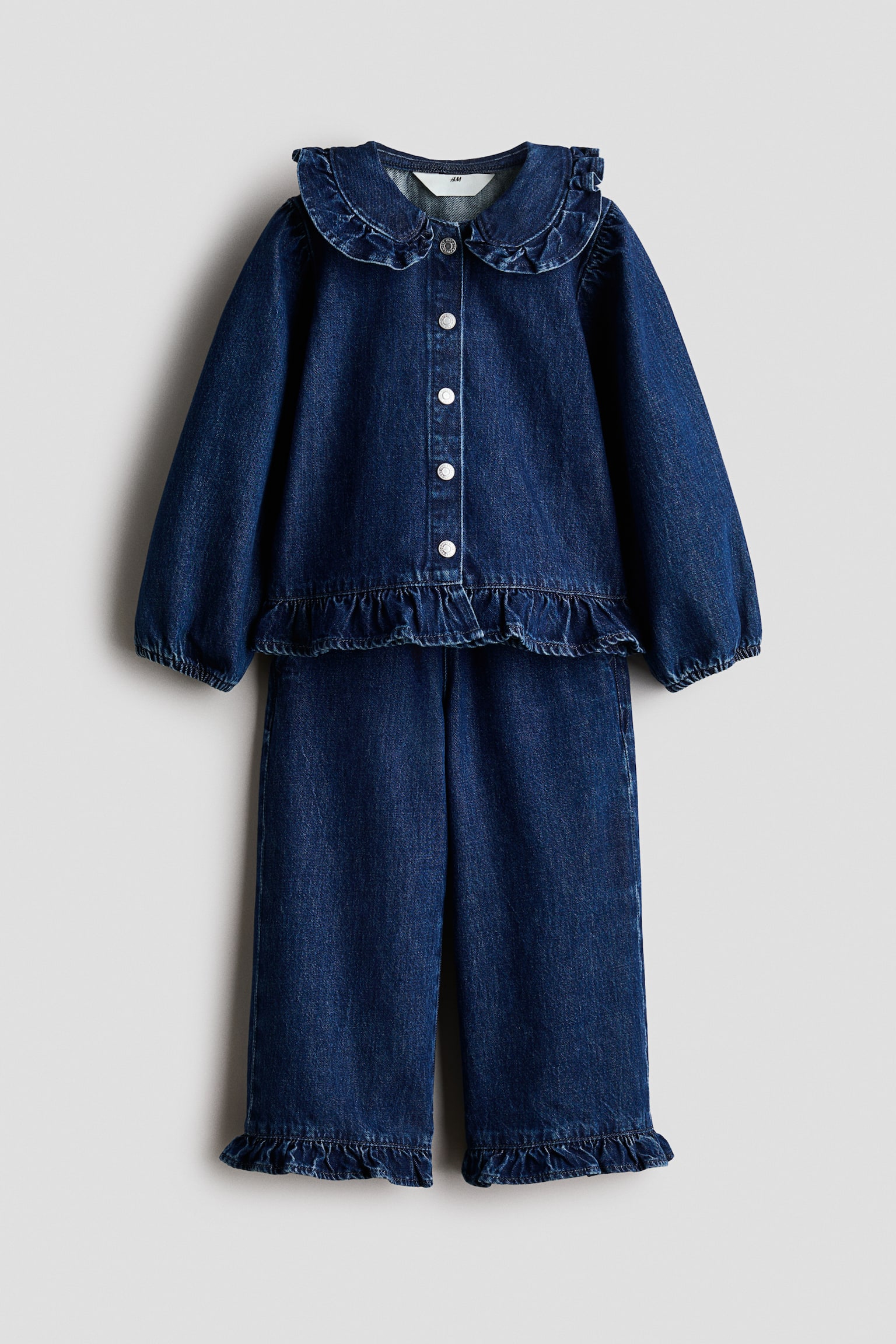 Een tweedelige donkerblauwe denimset wordt van voren getoond, bestaande uit een top met lange mouwen en een bijpassende broek. De top heeft een gerimpelde Peter Pan-kraag, een sluiting aan de voorkant met drukknoopjes en een gerimpelde zoom, terwijl de broek met wijde pijpen bij de enkels eveneens voorzien is van bijpassende gerimpelde zomen.