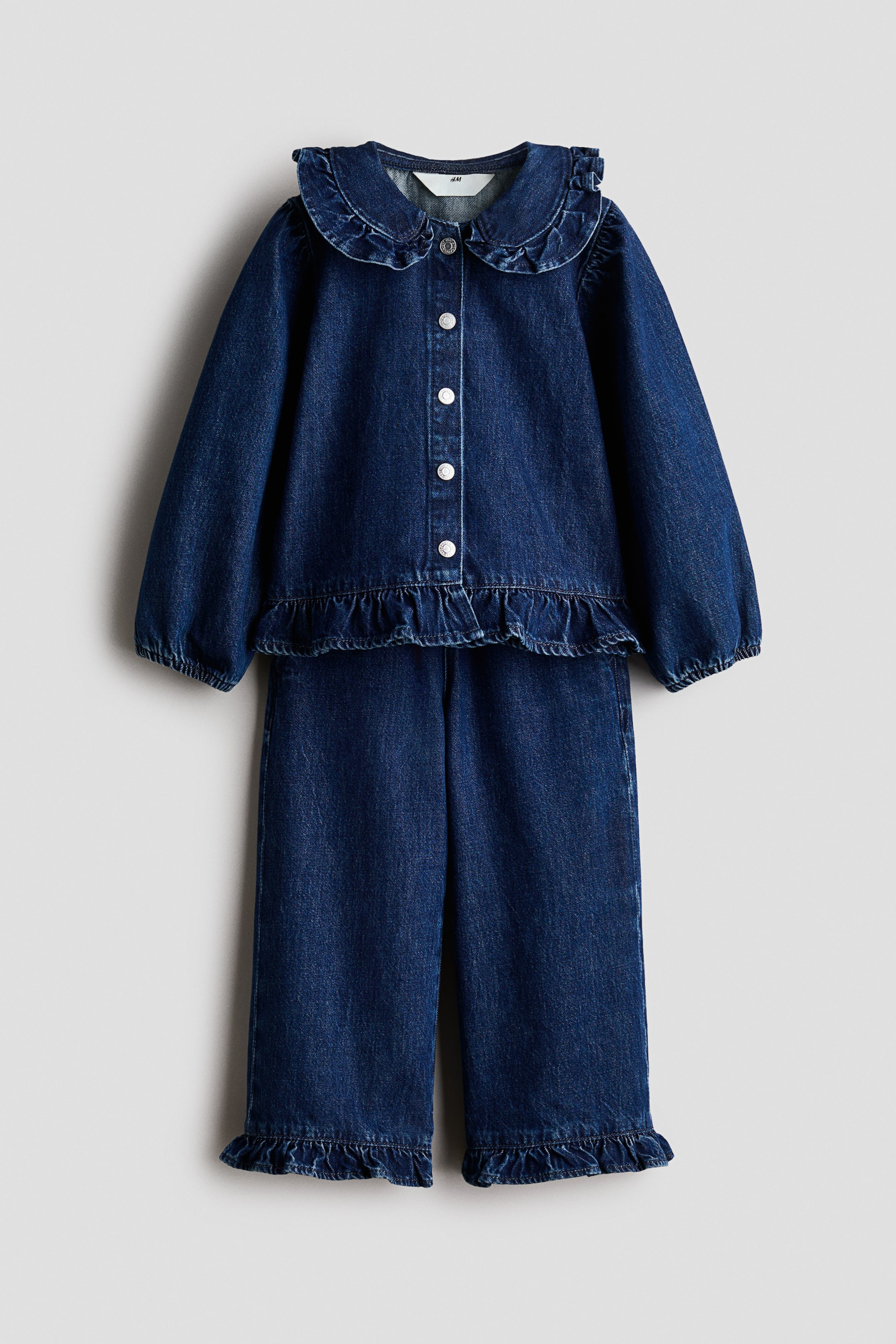 View larger image: A two-piece dark blue denim set is shown from the front, consisting of a long-sleeved top and matching pants. The top features a ruffled Peter Pan collar, a snap-button front closure, and a ruffled hem, while the wide-leg pants showcase matching ruffled hems at the ankles.