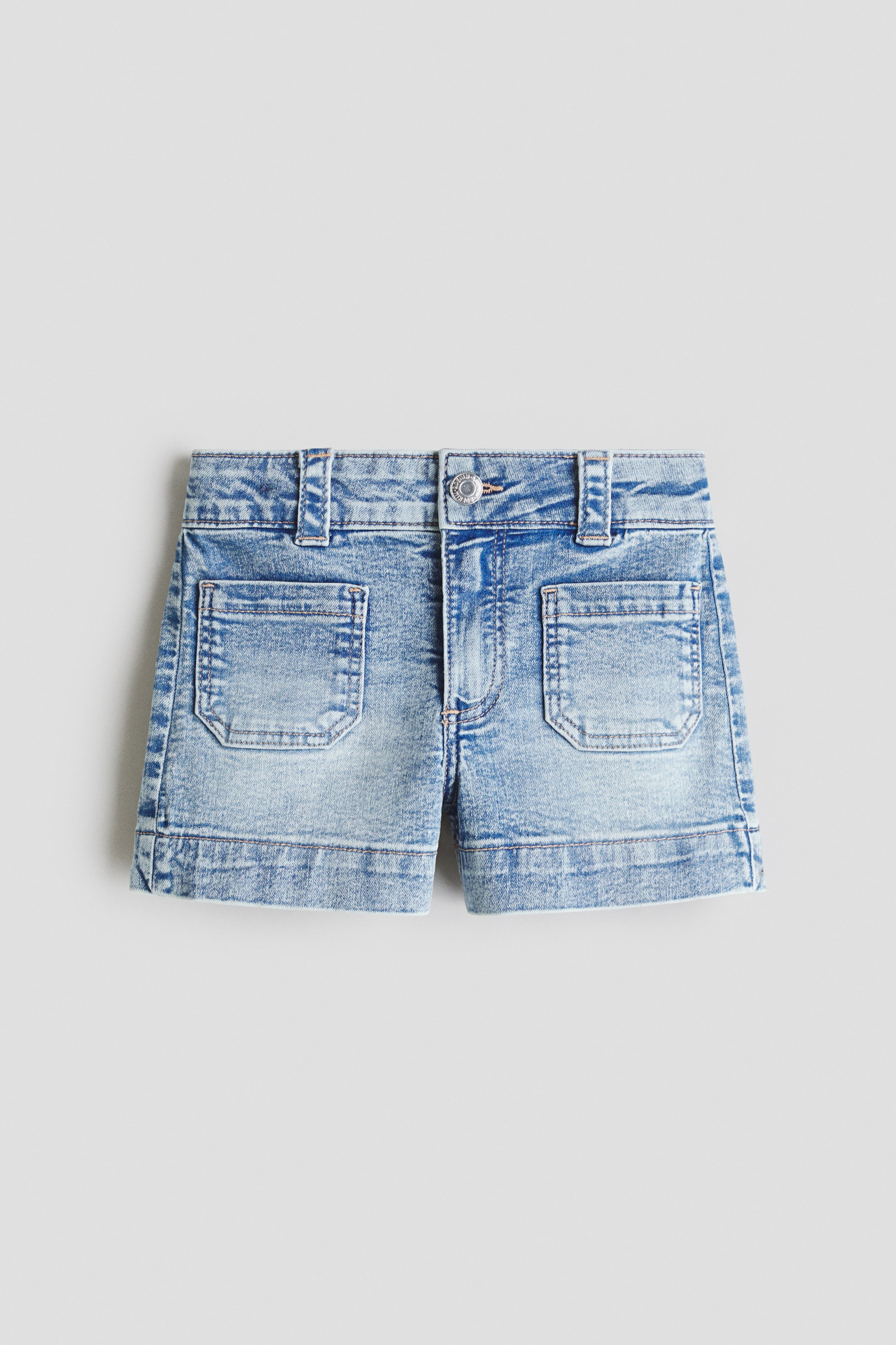 Größeres Bild ansehen: Jeansshorts zeigen ein hellblaues, acid-wash Finish mit einem einzelnen Knopfverschluss und Gürtelschlaufen am vorderen Bund. Zwei große, rechteckige Aufsatztaschen sind auf der Vorderseite sichtbar, eine auf jedem Bein, oberhalb der genähten, umgeschlagenen Säume.