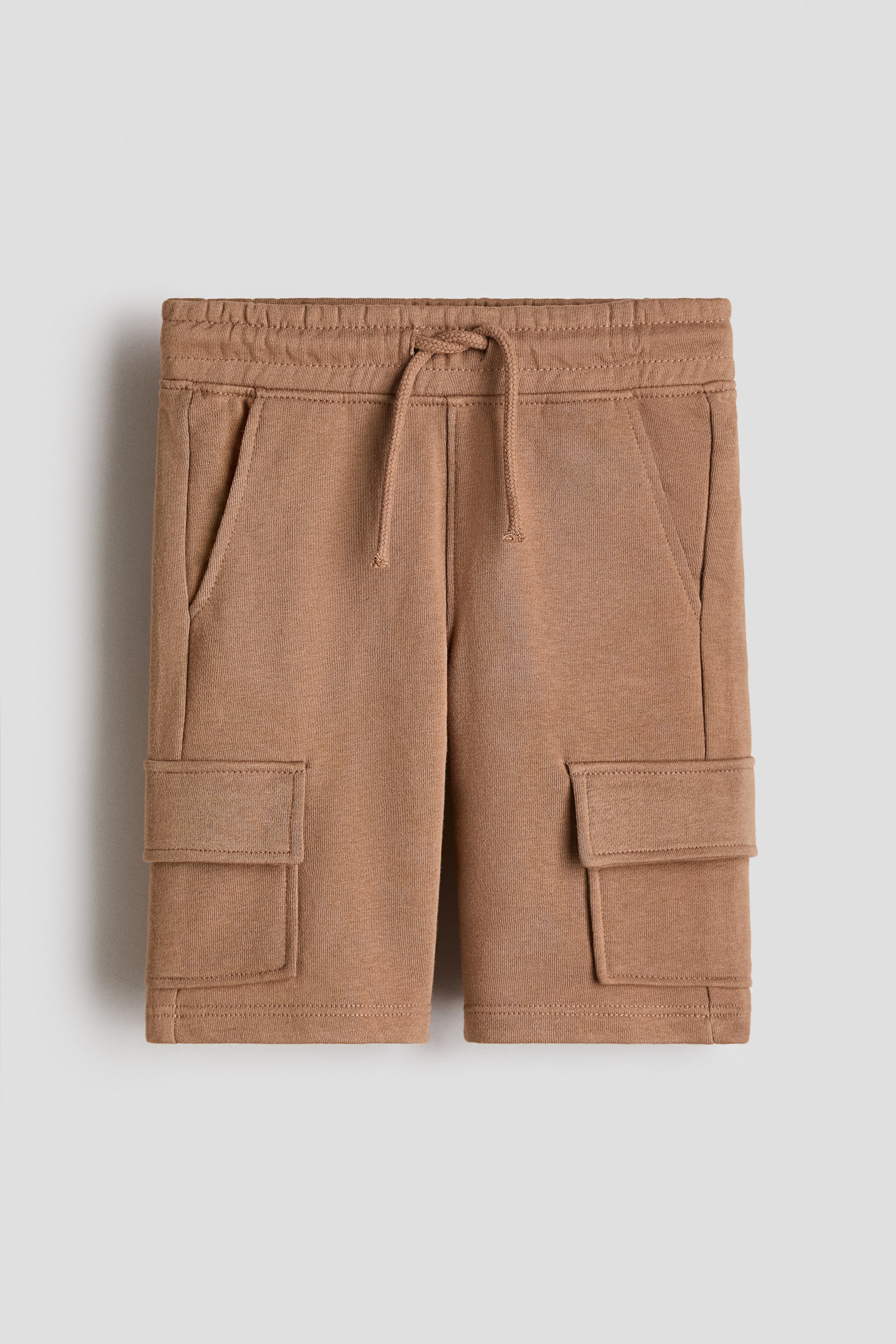 View larger image: The front of tan-colored sweatshorts is featuring an elasticized drawstring waistband, two angled side pockets, and two rectangular flap cargo pockets positioned on the outer thighs.