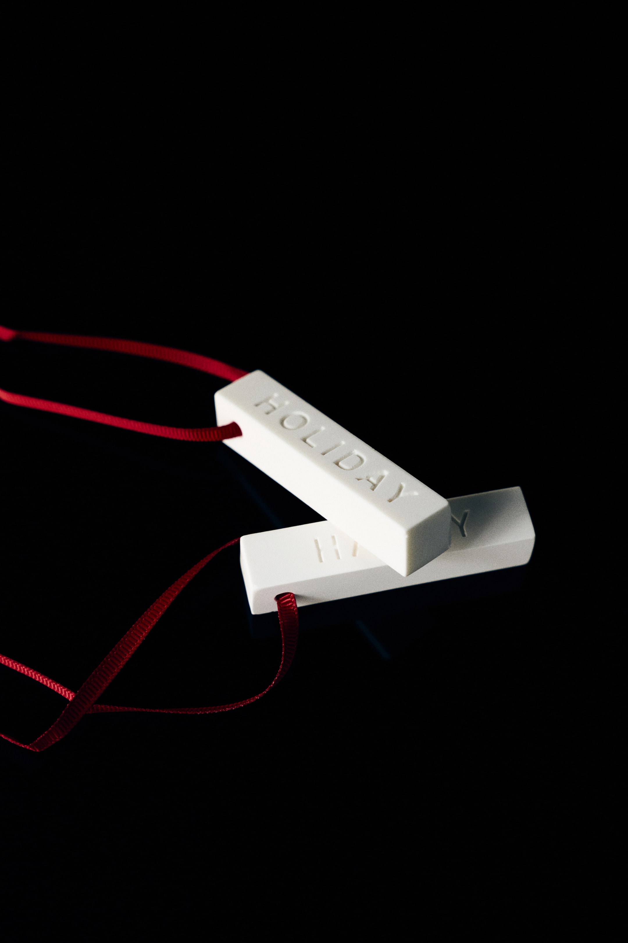 View larger image: Two white rectangular plaster bars are shown, with one resting diagonally atop the other. Each bar features the word "HOLIDAY" engraved on its surface and is threaded with a thin red grosgrain ribbon loop at one end.