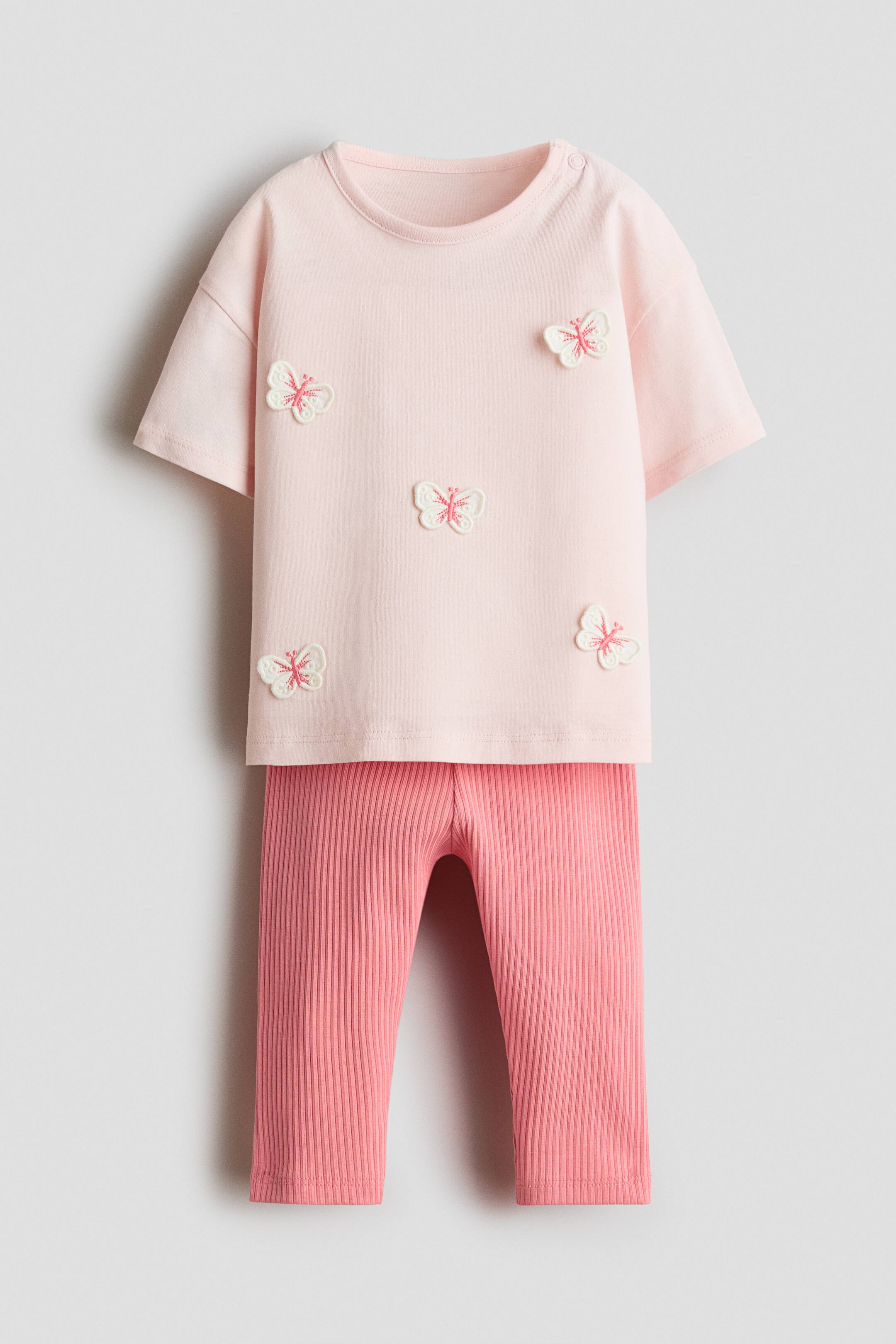 Xem ảnh lớn hơn: Một bộ cotton hai món được chụp từ phía trước, gồm áo thun tay ngắn màu hồng nhạt, được trang trí bằng các chi tiết đắp nổi hình bướm thêu màu trắng và hồng rải đều trên bề mặt vải. Áo thun được phối cùng quần legging gân đồng bộ với tông hồng đậm hơn.