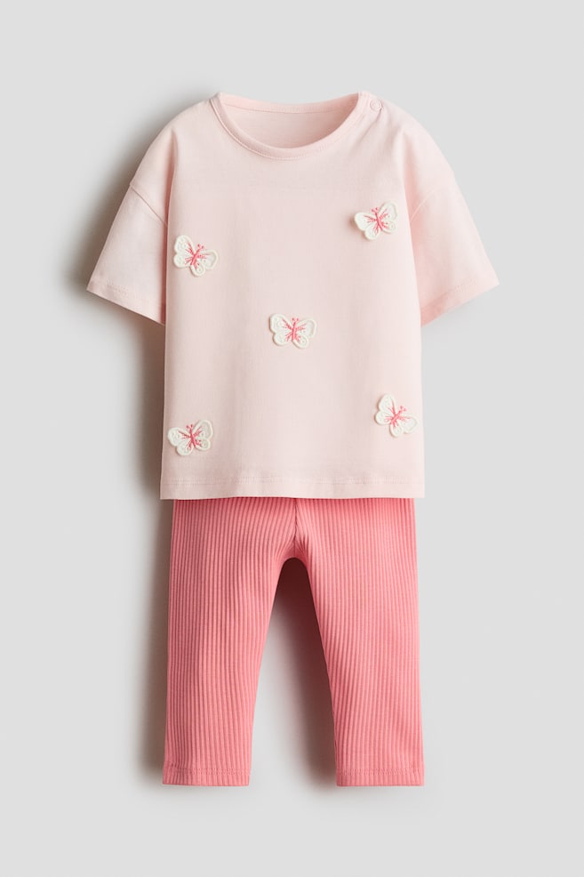 A two-piece cotton set is shown from the front, featuring a light pink, short-sleeved T-shirt decorated with embroidered white and pink butterfly appliquÃ©s scattered across the fabric. The T-shirt is paired with coordinating ribbed leggings in a darker shade of pink. - 1