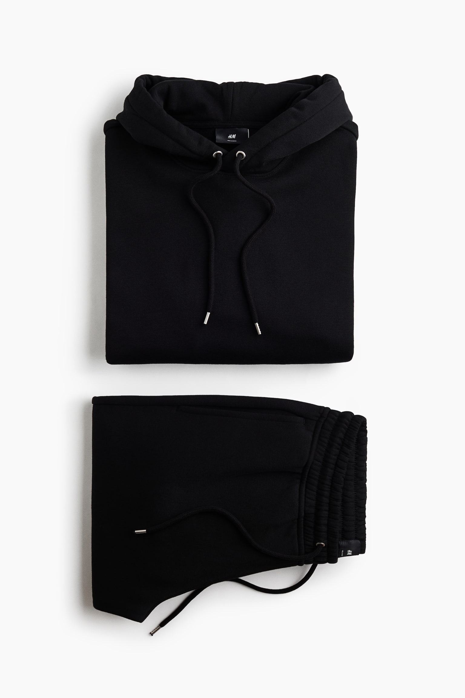 A folded black sweat fabric set is presented in a flat lay view, showing a hoodie positioned above matching track pants. The hoodie displays a drawstring hood with silver-tipped cords, while the track pants show an elasticated, smocked waistband with a matching drawstring.