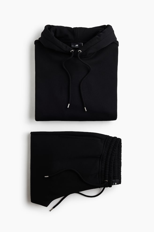 Ein zusammengefaltetes schwarzes Sweat-Set wird in einer Flatlay-Ansicht präsentiert, wobei ein Hoodie über einer passenden Jogginghose angeordnet ist. Der Hoodie zeigt eine Kapuze mit Kordelzug und silberfarbenen Kordelenden, während die Jogginghose einen elastischen, gesmokten Bund mit passendem Kordelzug aufweist. - 1