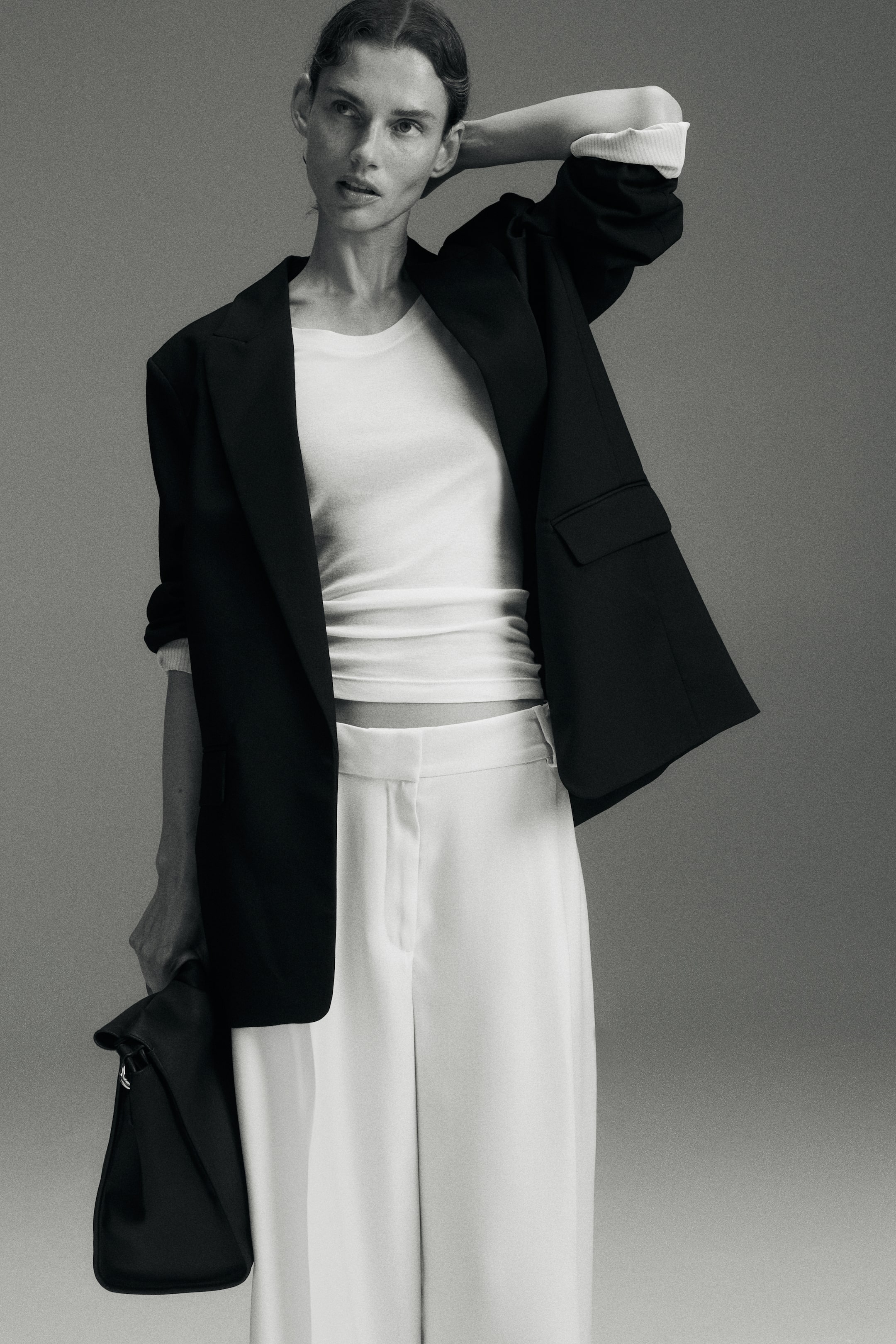 View larger image: An oversized, long black blazer in a woven fabric is worn open at the front, layered over a white T-shirt and white trousers. The blazer features structured shoulder pads, peak lapels, and a jetted pocket with a flap, showcasing its relaxed, fastening-free design.