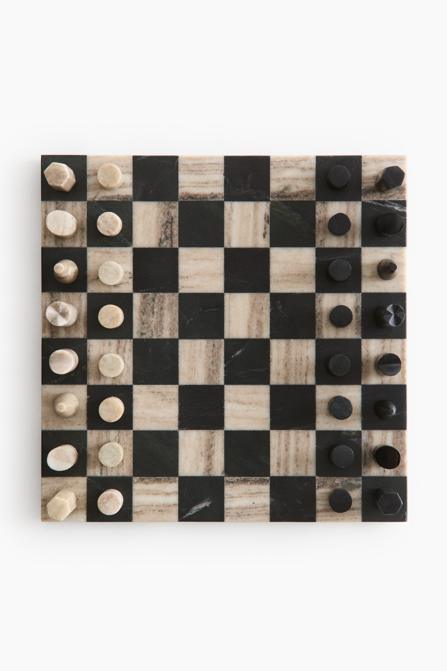 A square stone chessboard displays alternating dark grey and beige veined squares, seen from a direct overhead angle. Minimalist, geometric chess pieces made of matching light beige and dark grey stone stand in their starting positions on opposite sides of the board.