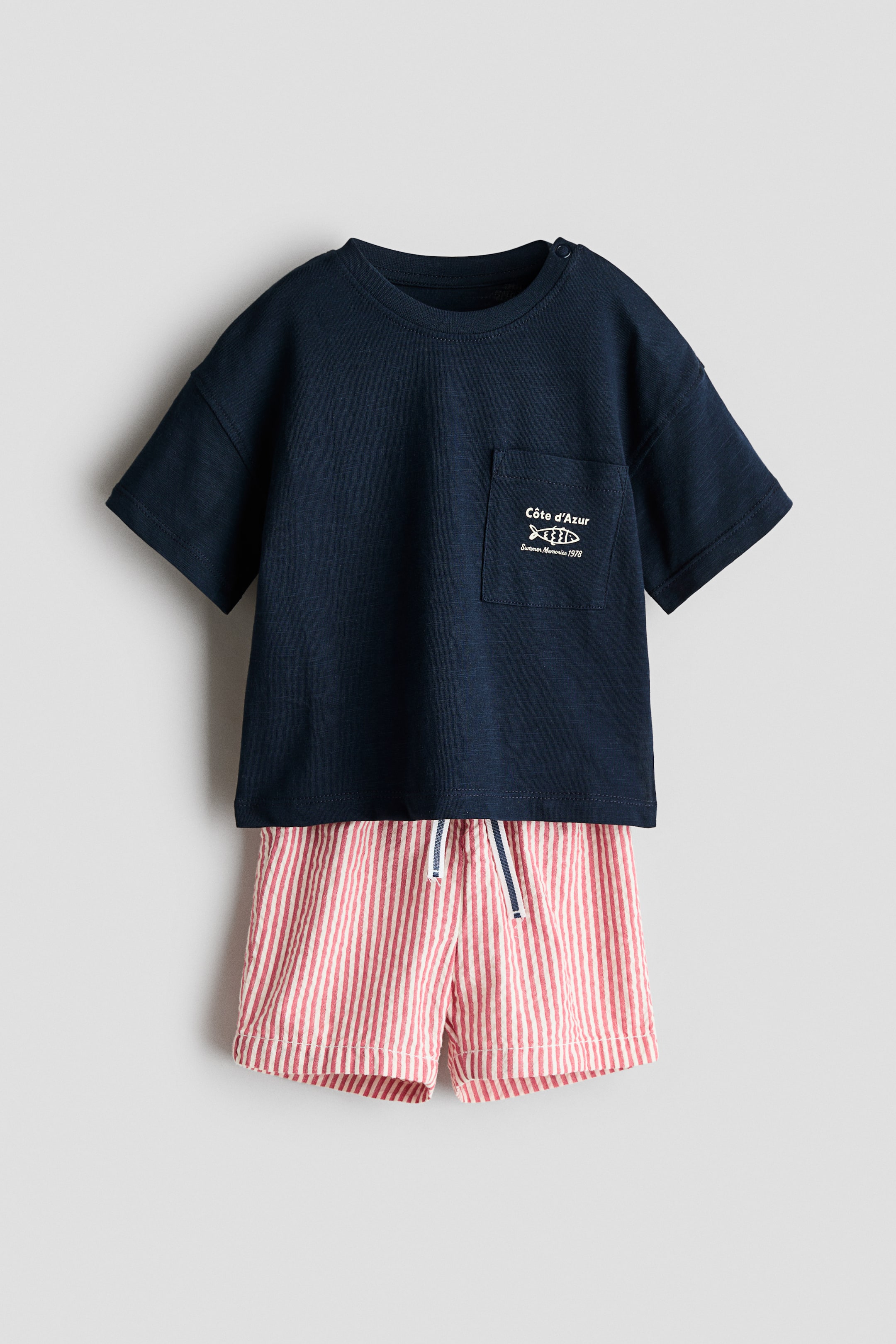 View larger image: A baby's two-piece outfit is presenting, featuring a navy blue t-shirt and striped shorts. The t-shirt is displaying short sleeves, a round neck with shoulder snaps, and a left chest pocket with a white "Côte d'Azur" fish graphic. The shorts are showcasing red and white vertical seersucker stripes and an elastic waistband with a navy and white striped drawstring.