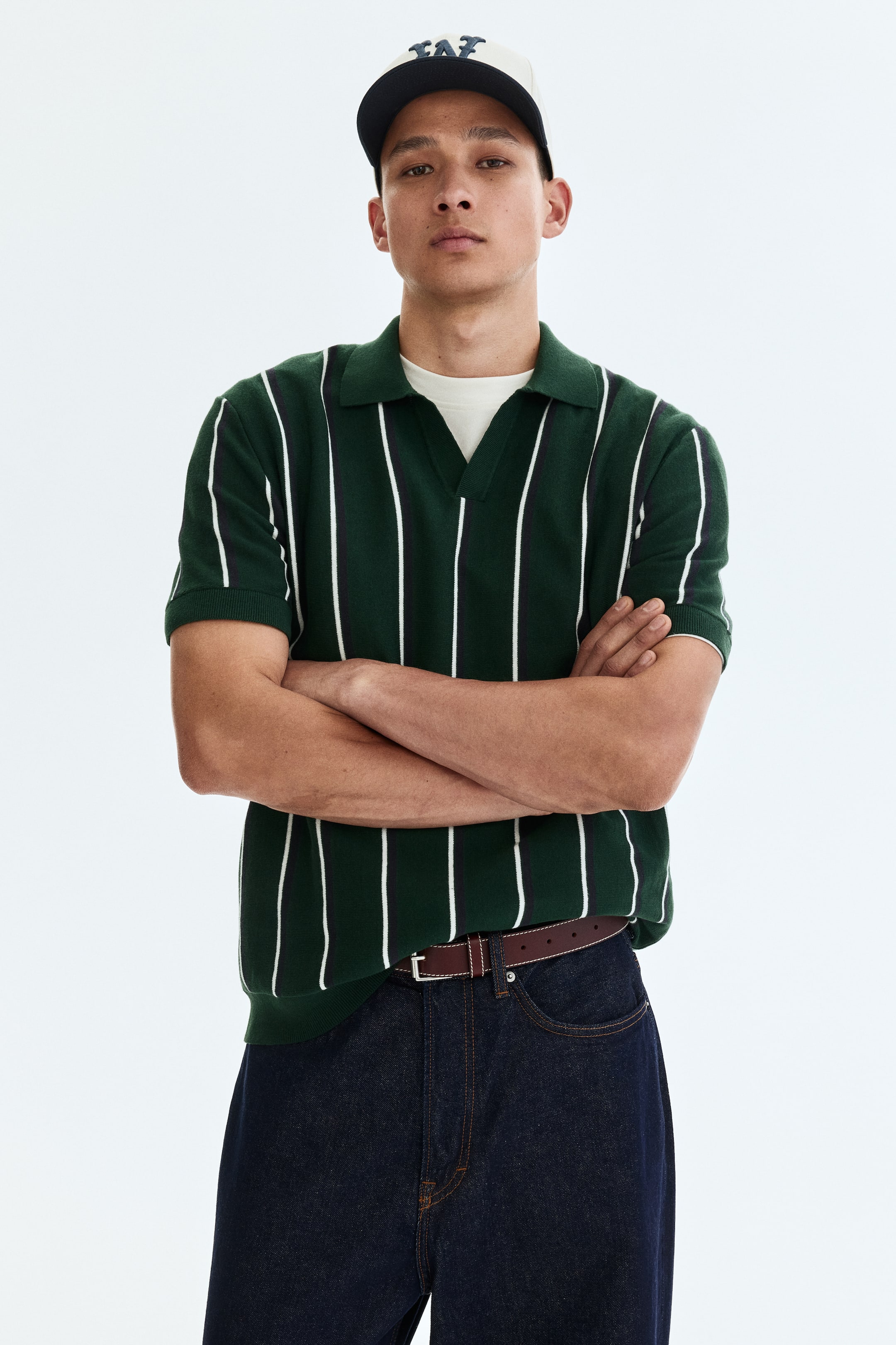 View larger image: The model is wearing a short-sleeved polo shirt crafted from a dark green knit with white vertical stripes. It showcases a ribbed collar, a V-shaped front opening, and a straight hem, layered over a light-colored t-shirt.