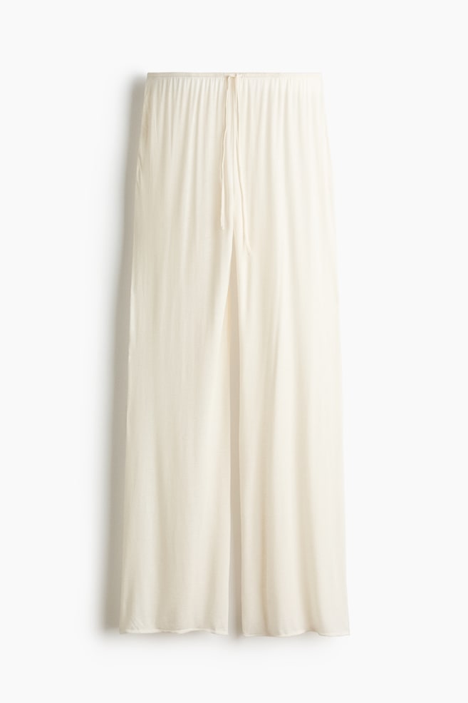 These cream-coloured, wide-leg trousers are hanging front-facing, showcasing a gathered elastic waistband with a thin drawstring tie at the centre. The lightweight, fluid fabric drapes down to form a relaxed, loose-fitting silhouette. - 1