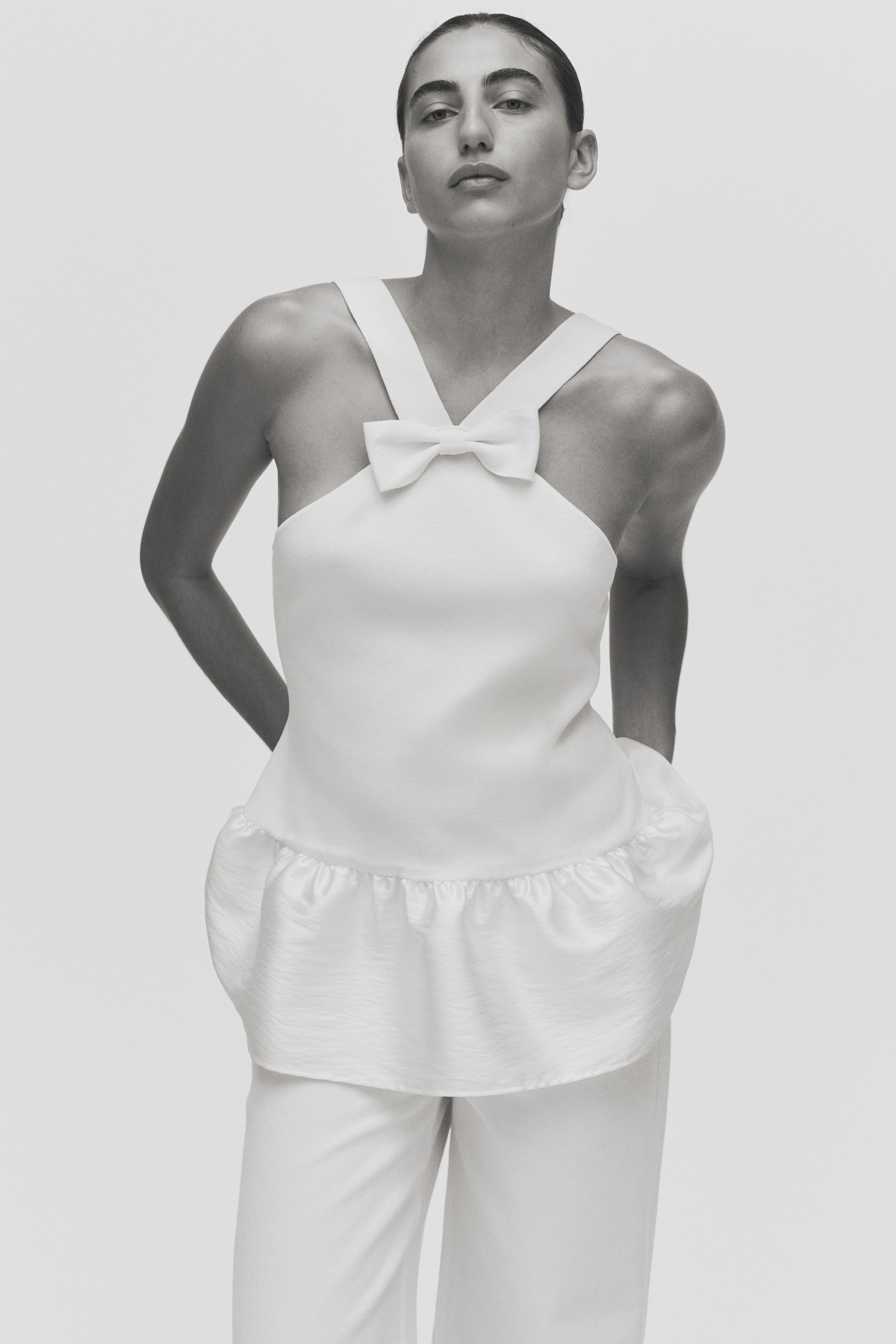 View larger image: A white two-piece ensemble is being presented from the front, comprising a halter-style top and coordinating relaxed-fit pants. The top features wide shoulder straps converging at a small bow at the center neckline, and a ruffled peplum hem, with both pieces showcasing a subtle textured fabric.