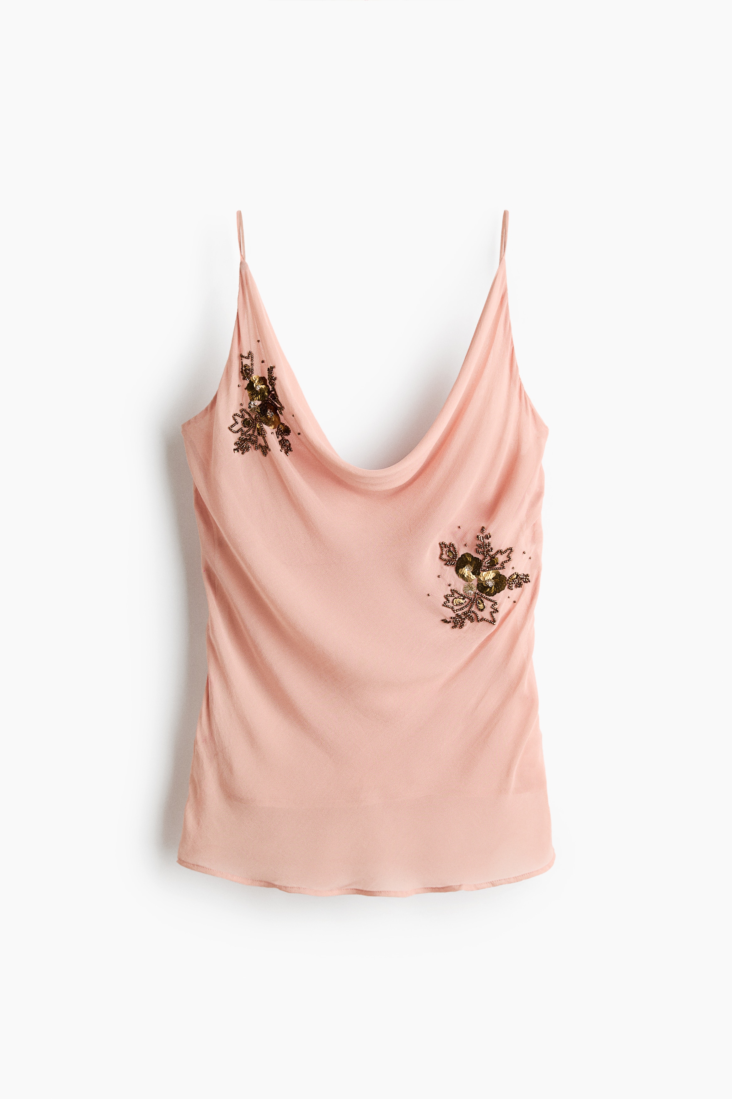 The model is wearing a light pink top featuring a draped cowl neckline, thin spaghetti shoulder straps, and a beaded floral embellishment on the front. She is posing with one arm crossed over her waist, showcasing the topâs lightweight, fluid fabric.