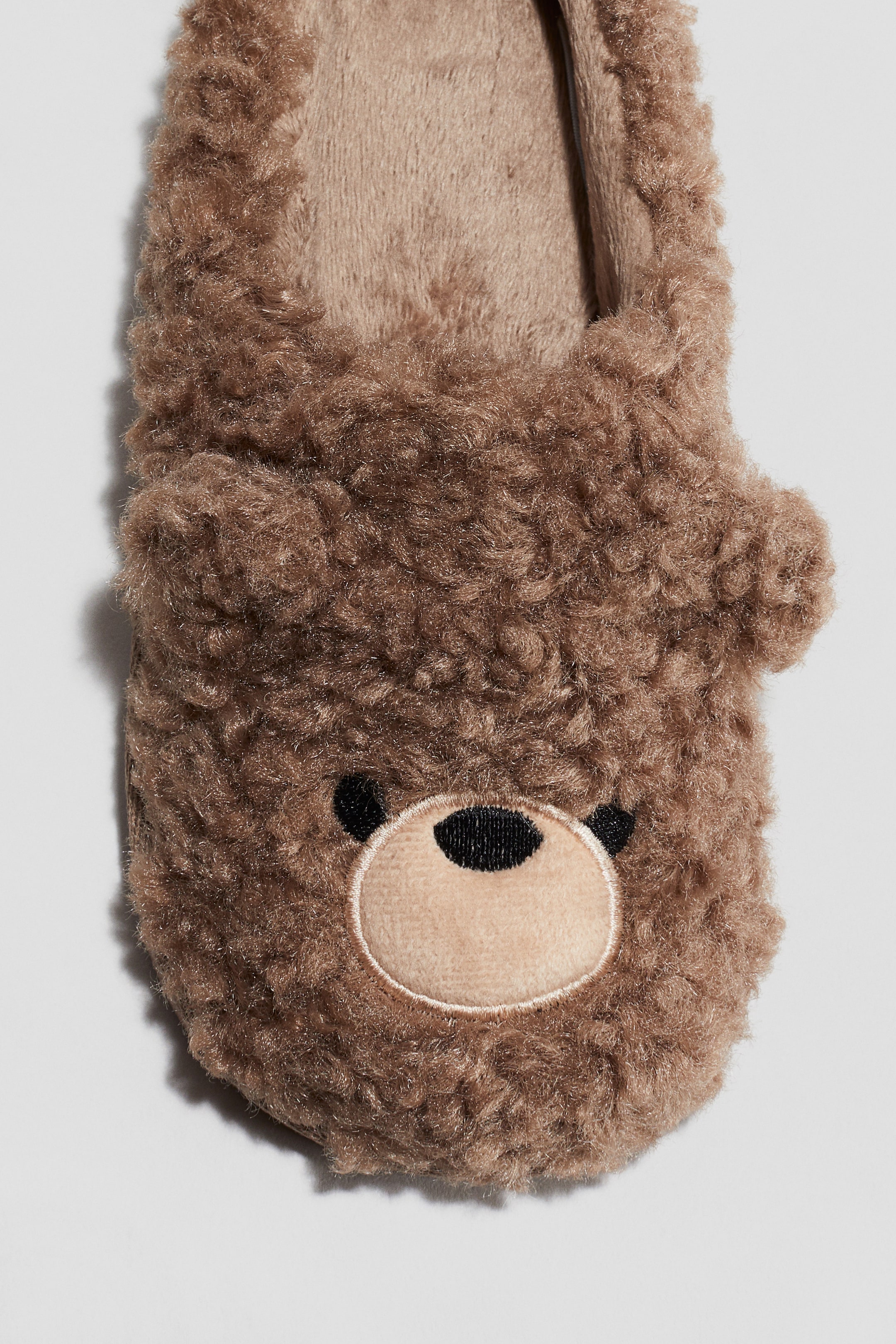 Grotere afbeelding bekijken: Een bruine, pluizige pantoffel staat naar voren gericht, met een geborduurd berengezicht op de bovenkant. Het gezicht bevat een lichtere, ronde snuit met een zwart geborduurde neus en twee zwarte ogen, en kleine ronde oren zijn zichtbaar in de bovenhoeken.