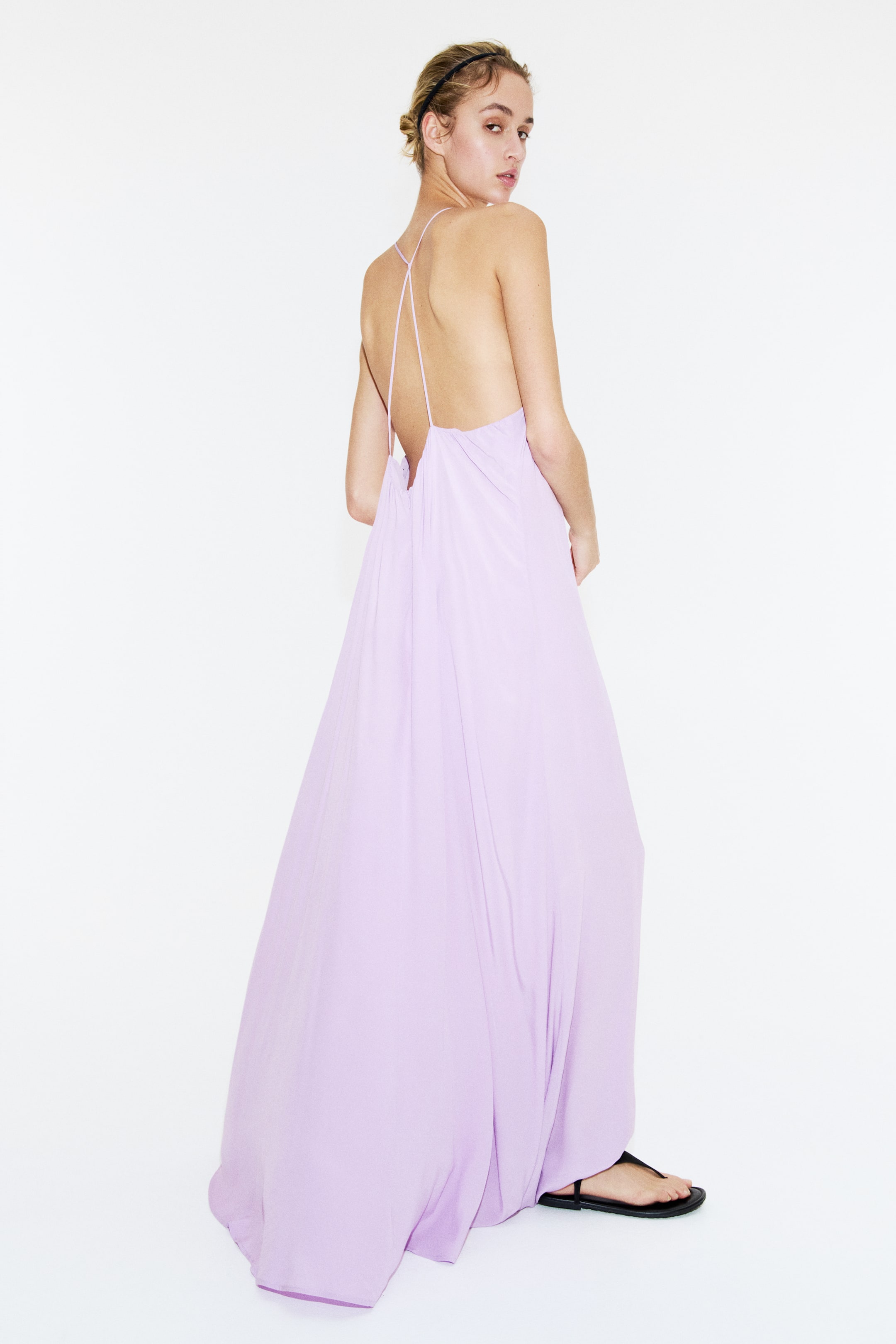 View larger image: A lavender maxi dress is showcased from a back-three-quarter angle, highlighting a low open back with delicate crisscross straps. The flowing fabric drapes loosely to the floor.