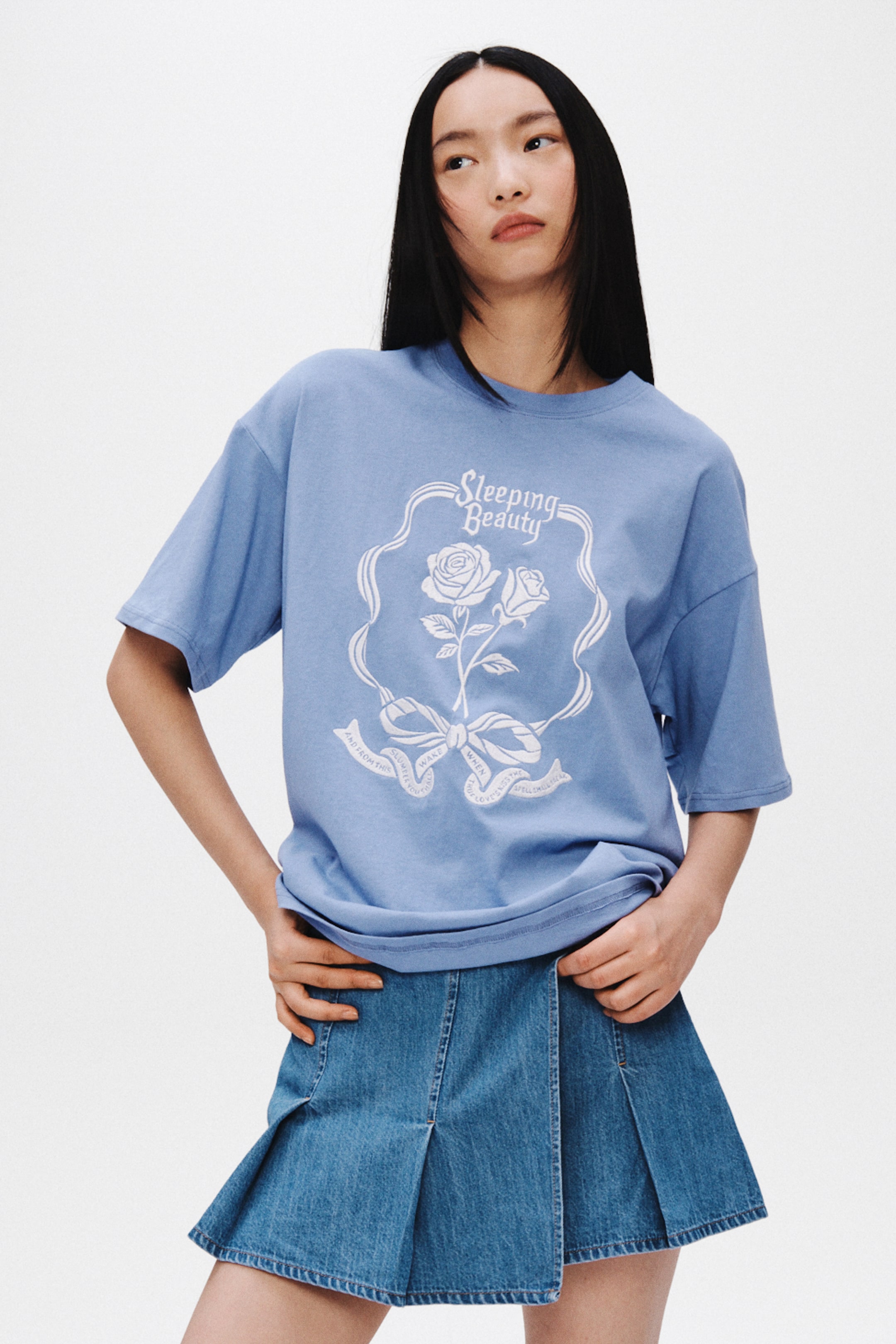 Xem ảnh lớn hơn: Chiếc áo phông oversized màu xanh nhạt có một họa tiết in lớn màu trắng ở mặt trước, với dòng chữ "Sleeping Beauty" nằm phía trên hai bông hồng, một chiếc nơ và một dải băng có chữ, tất cả được bao bọc trong một khung trang trí. Áo được sơ vin lơi vào chân váy denim, phô bày phom dáng rộng rãi, thoải mái.
