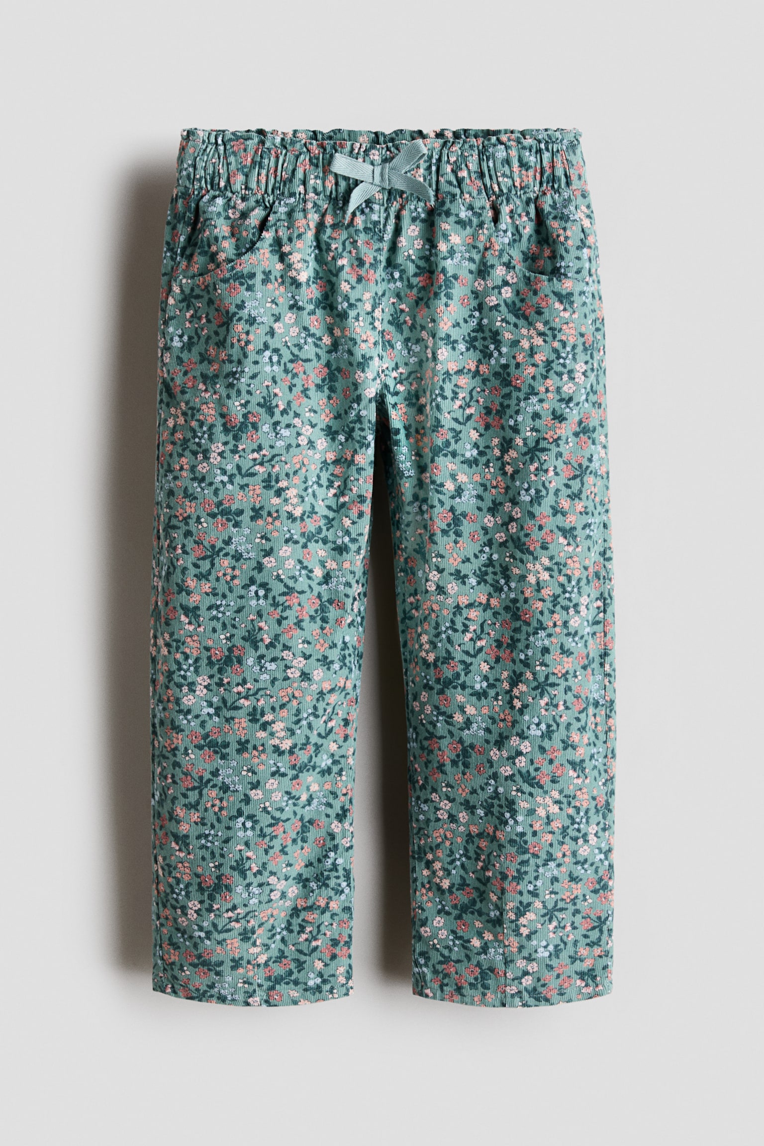 Een corduroy broek wordt van voren getoond, met een all-over ditsy bloemenpatroon in roze, wit en blauw op een saliegroene achtergrond. De broek is ontworpen met een elastische tailleband, een bijpassend trekkoord aan de voorkant in strikvorm, zijzakken en een rechte pijpsilhouet.
