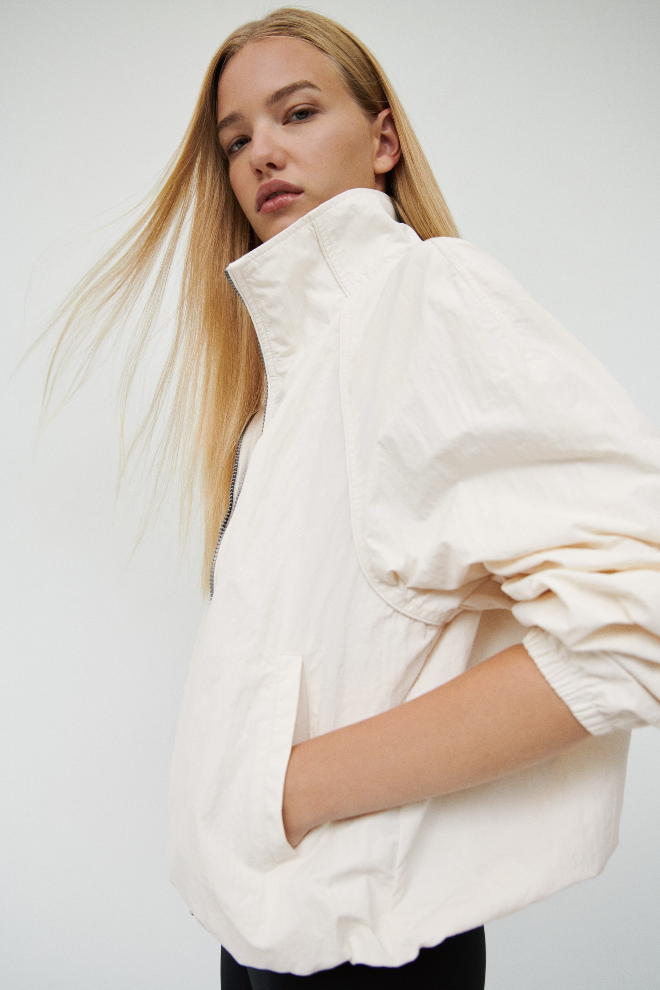 Voir une image plus grande: Une veste tissée surdimensionnée blanc cassé est présentée de profil droit, avec un col montant haut, de longues manches raglan à poignets élastiqués et un bas bouffant élastiqué. Le modèle repose sa main dans une poche passepoilée diagonale à l’avant, mettant en valeur la silhouette décontractée et volumineuse de la veste.