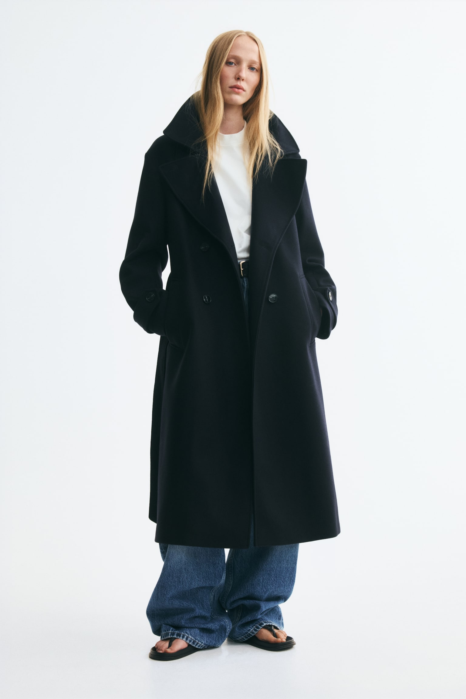 A dark navy double-breasted maxi coat is hanging front-facing, featuring a high funnel collar with a buttoned throat latch and a matching tie belt knotted at the waist. The coat is designed with dropped shoulders, long sleeves with buttoned cuff tabs, and subtle front welt pockets. - 1