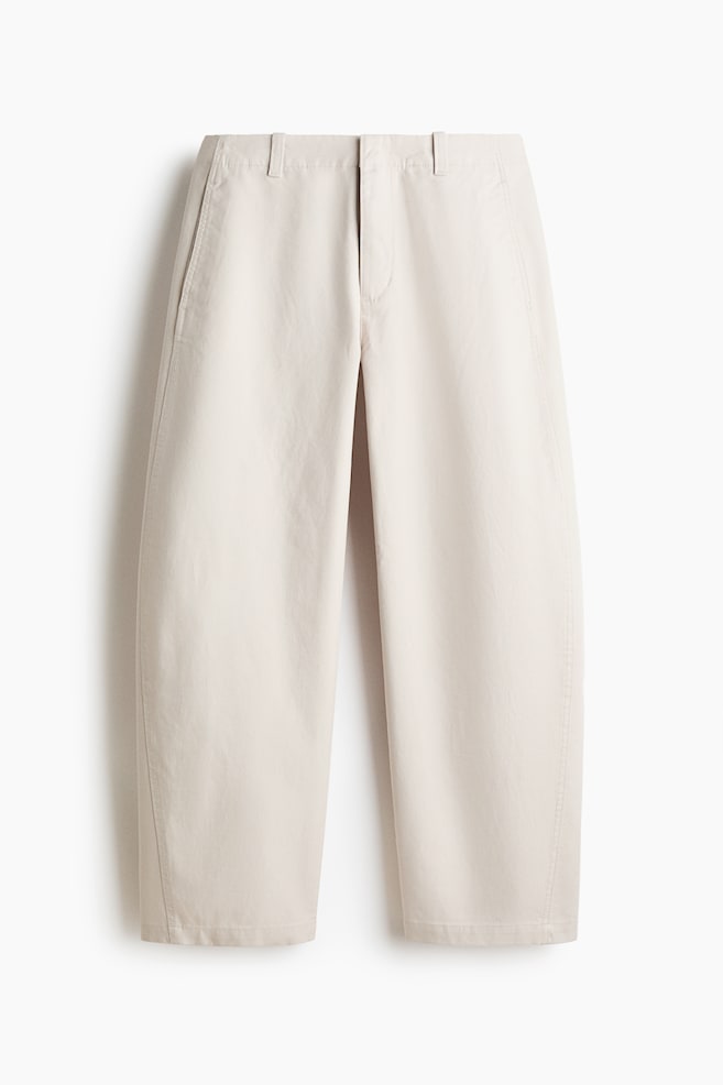 Die hellbeigen Hosen sind nach vorne gerichtet und verfügen über einen flachen Bund mit Gürtelschlaufen und seitlichen Eingrifftaschen. Sie präsentieren eine weite, leicht fassförmige Beinsilhouette mit vertikal gesteppten Nähten, die an jedem Bein bis zu einem verkürzten Saum verlaufen. - 1