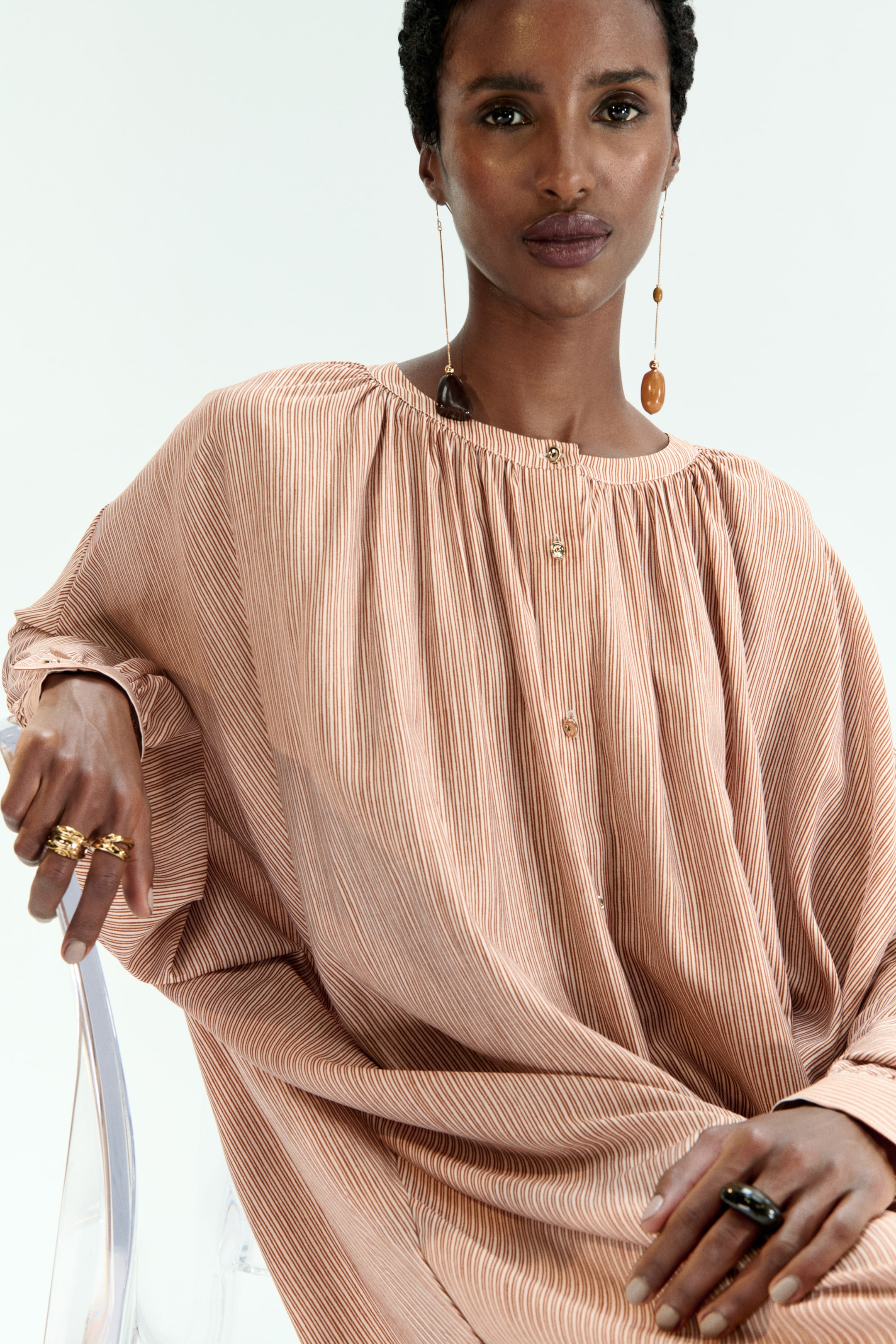 View larger image: A loose-fitting blouse in light peach with fine vertical brown stripes is featuring a gathered round neckline, a partial front button placket, and long, voluminous sleeves with buttoned cuffs, draping fluidly over the body.